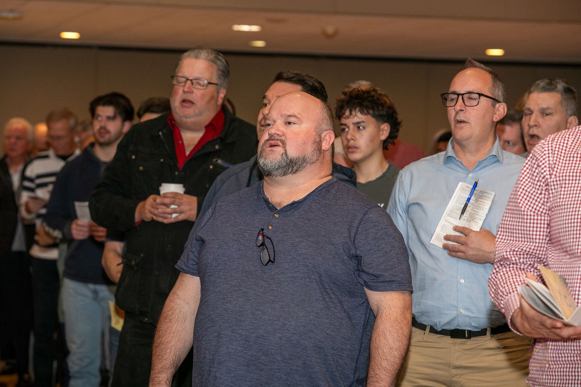 People standing indoors at a meeting, several holding drinks and papers, listening attentively