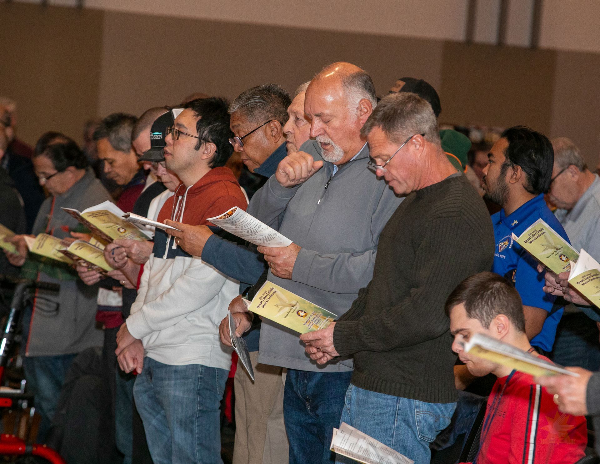 Audience members standing and reading booklets at a Catholic men's conference.