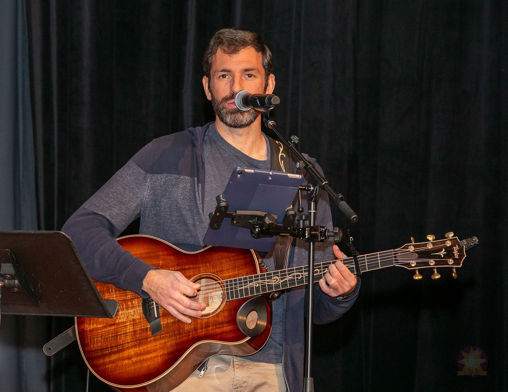 Musician singing into a microphone while playing acoustic guitar on stage with a music stand nearby