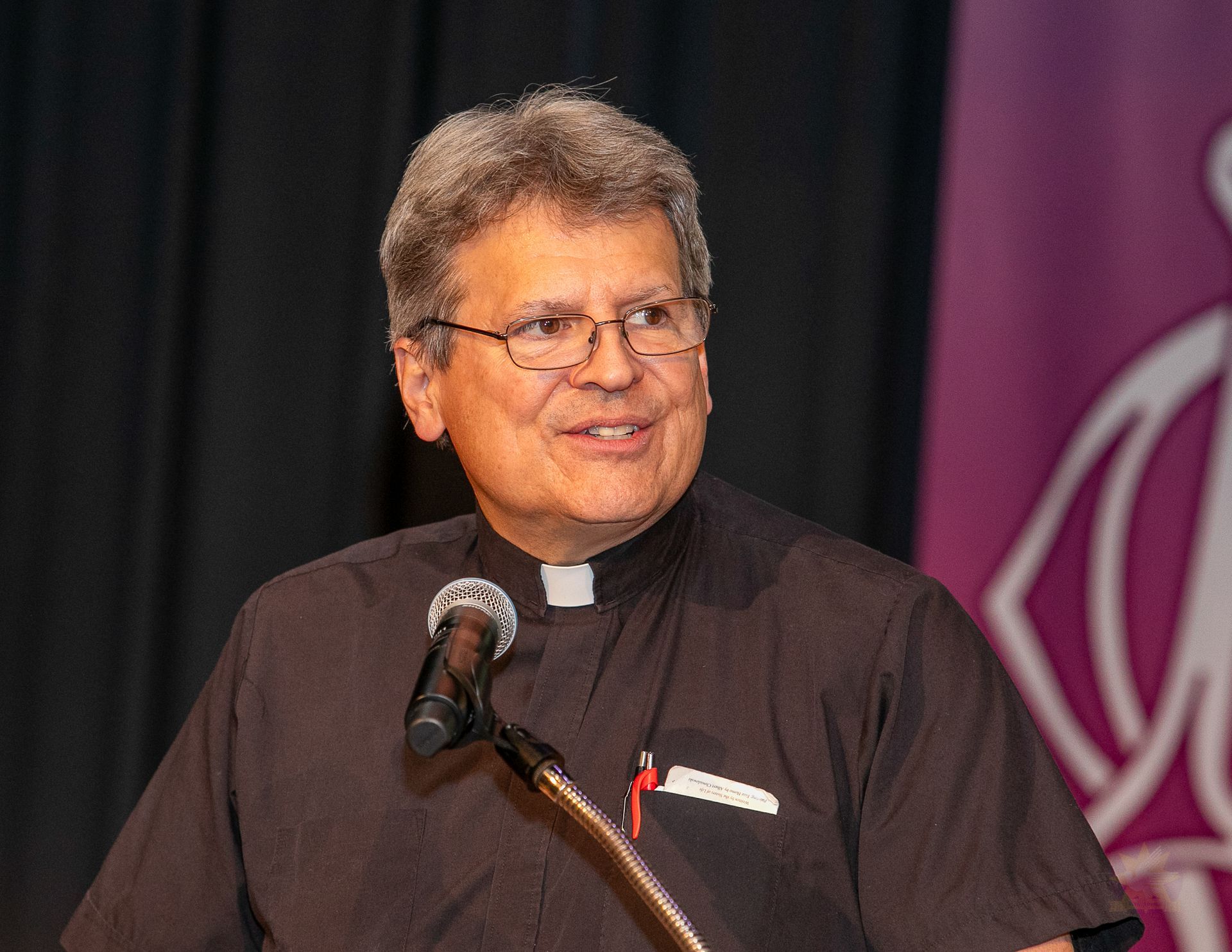 Priest speaking at a podium with a microphone, wearing a black clerical shirt and smiling onstage