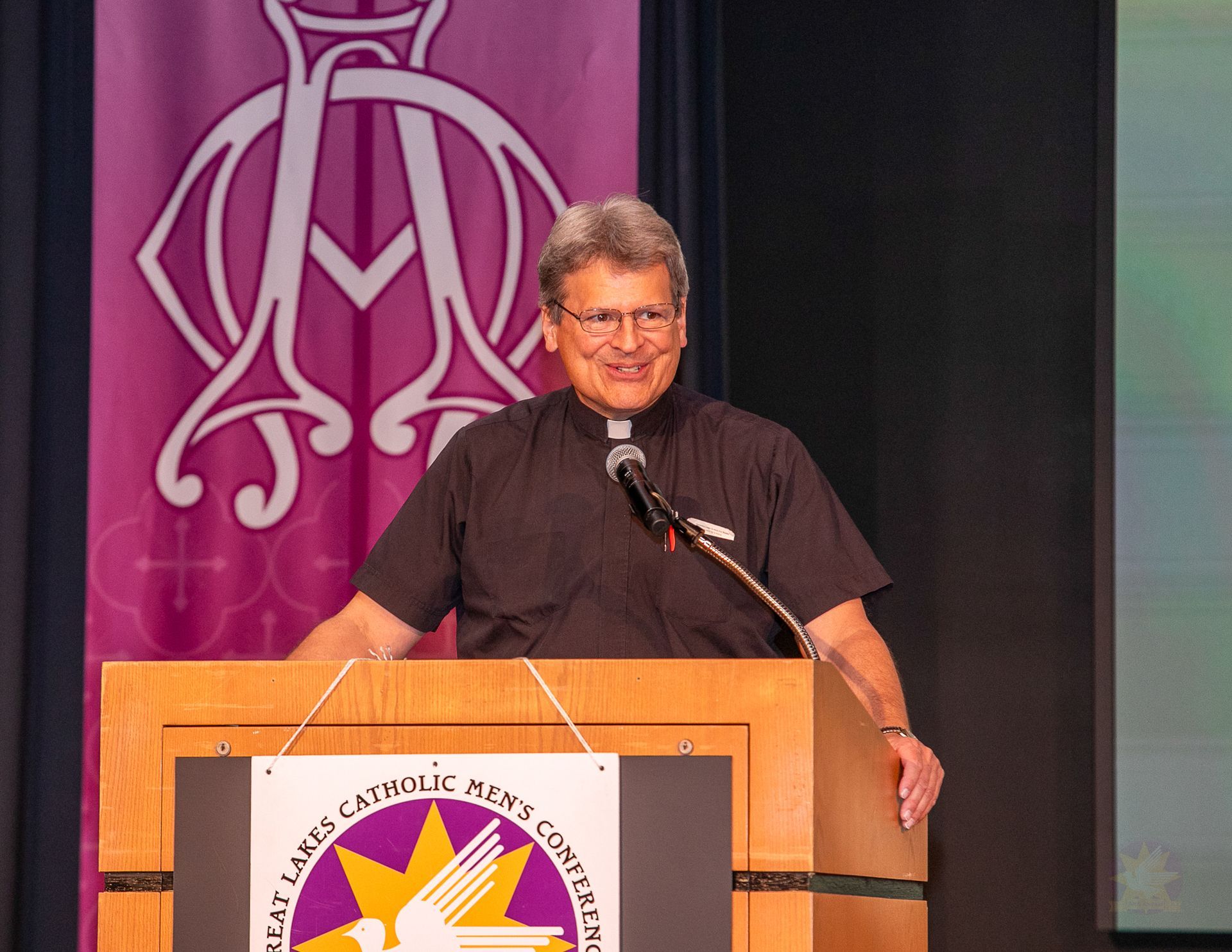 Priest speaking at a podium with a microphone on a stage, in front of a purple banner and black curtain.