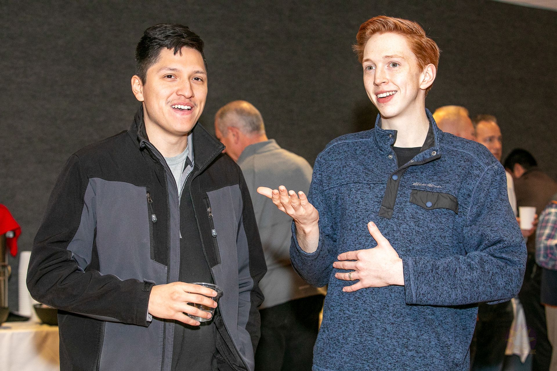 Two people chatting and smiling at an indoor event, one in a dark jacket and one in a blue jacket.