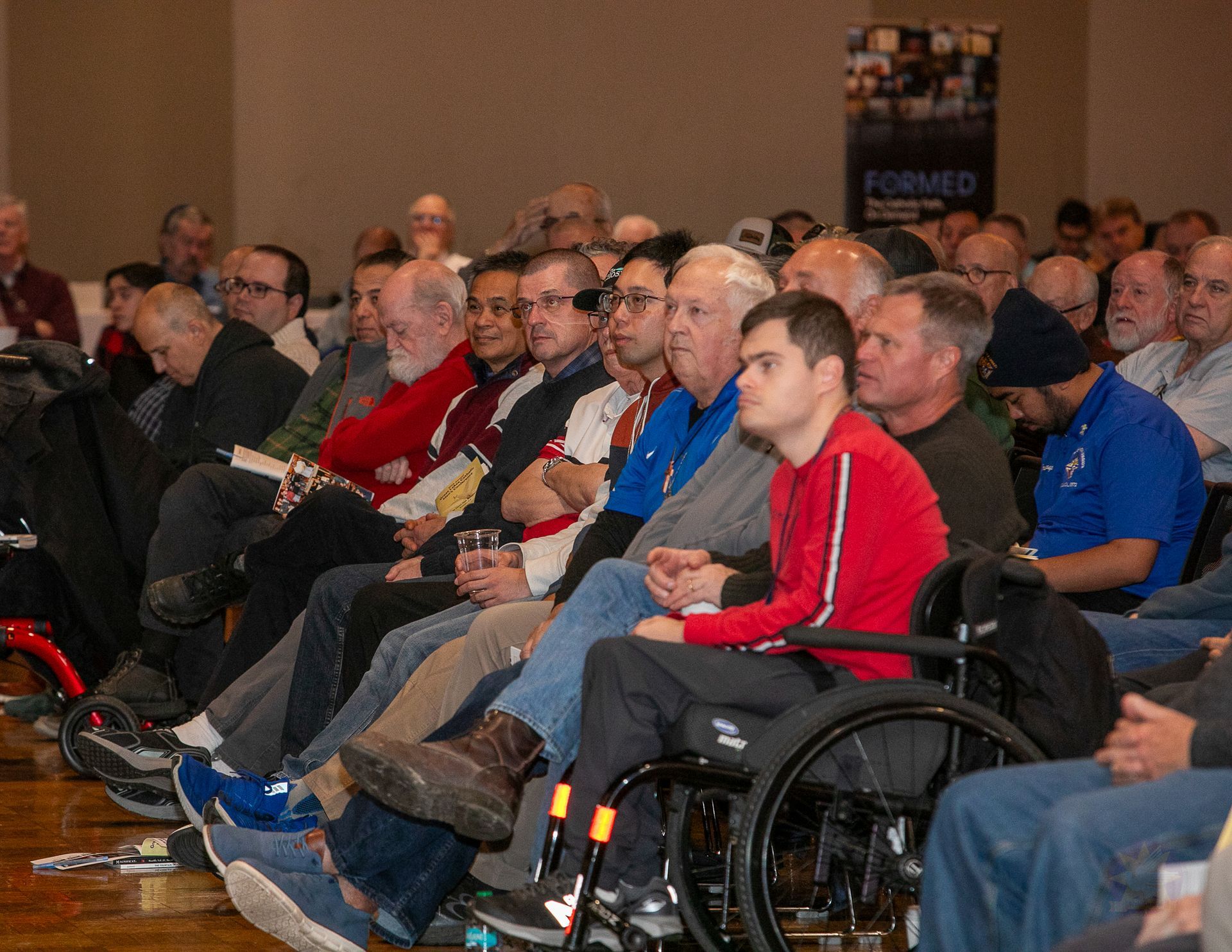 Audience seated in a conference room, many in wheelchairs, listening attentively during a presentation
