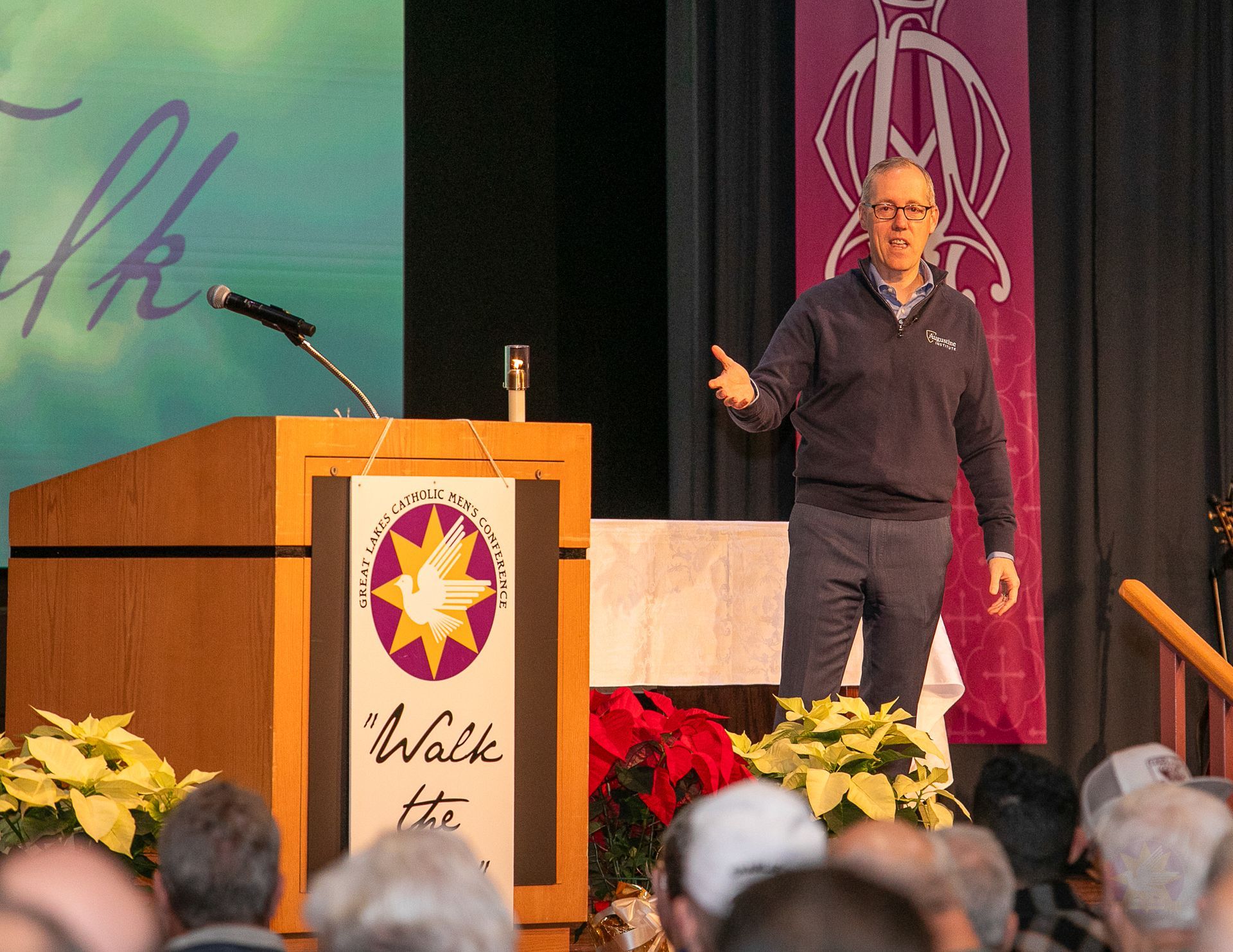 Dr. Tim Gray on stage beside podium with purple banner, addressing an audience at a Catholic men's conference.