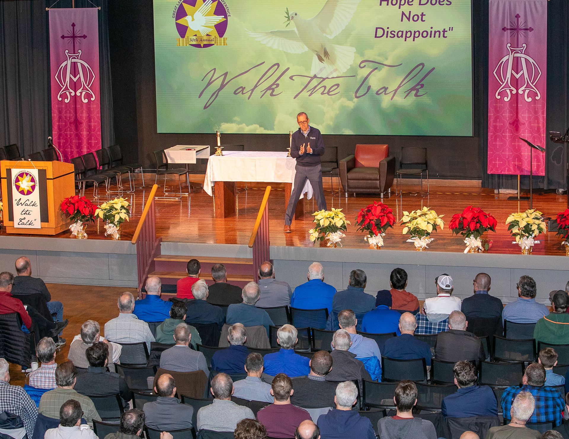 Speaker on church stage addressing seated audience beneath “Walk the Talk” banner.