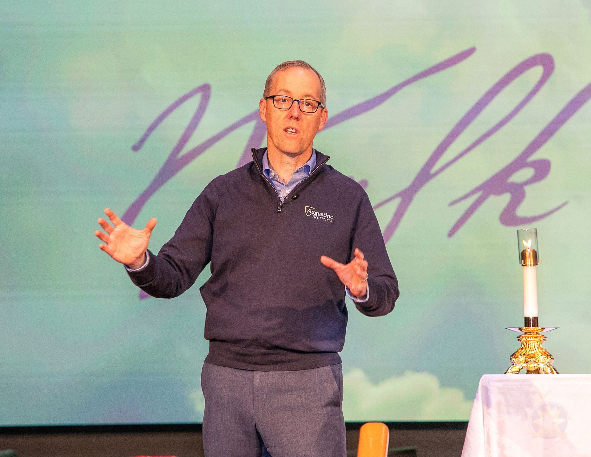 Man speaking onstage beside a table with a lamp, in front of a large purple script backdrop.