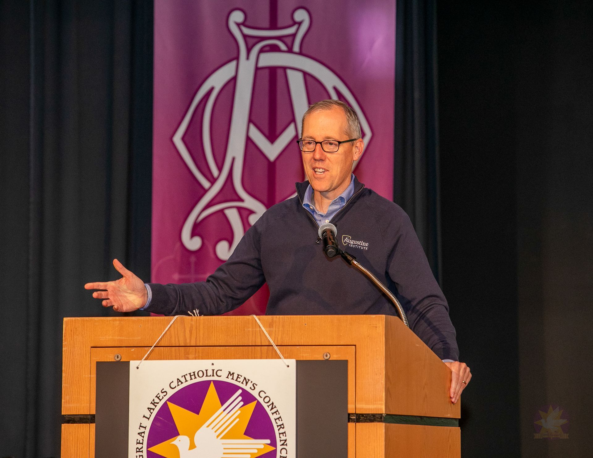 Dr. Tim Gray at the podium on stage, gesturing with one hand against a dark backdrop with a purple logo.