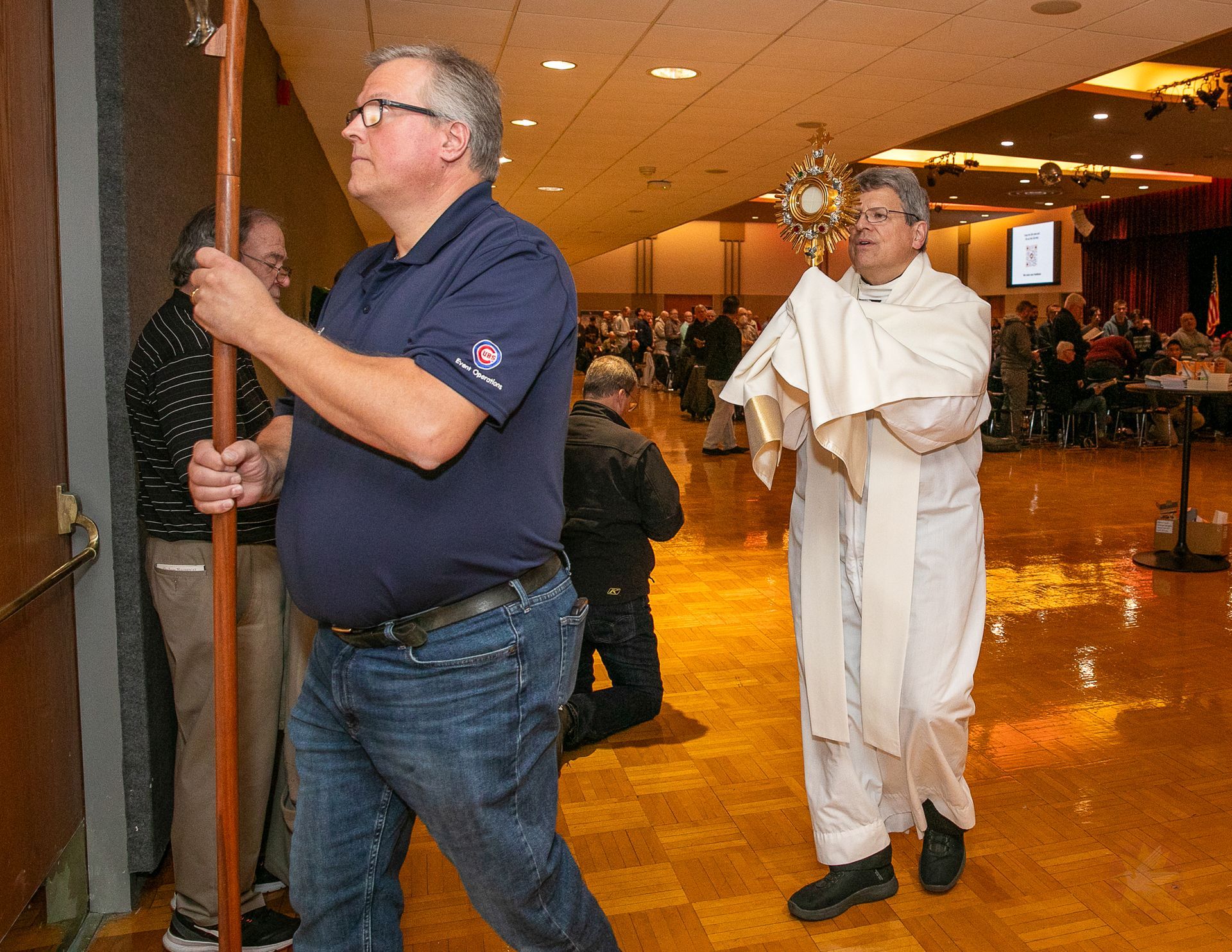 Man in navy shirt carries a crucifix while a priest carries a monstrance.