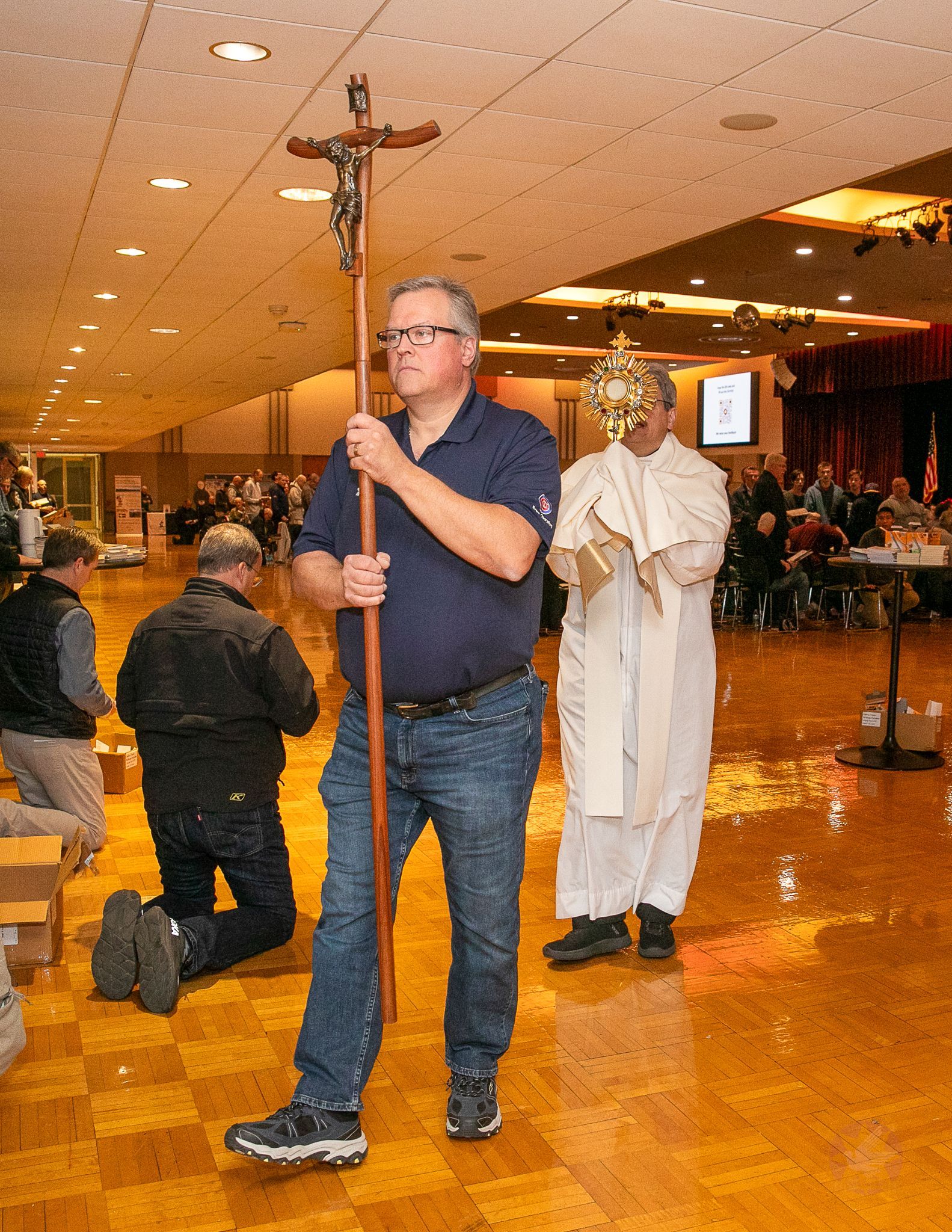 Man carrying a crucifix in a church hall, with a priest carrying a monstrance behind him.