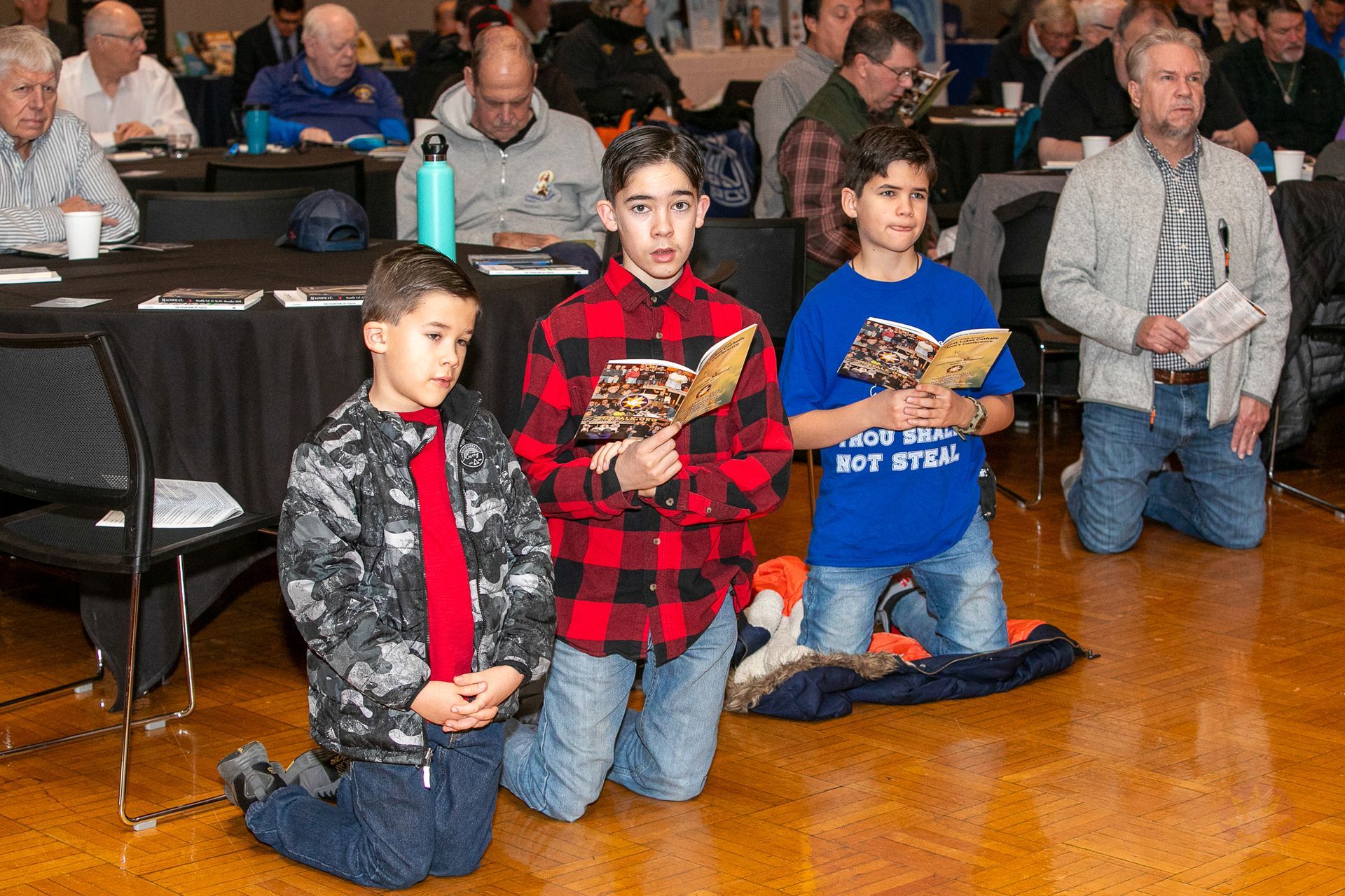 Three children kneeling indoors with adults behind them during a ceremony
