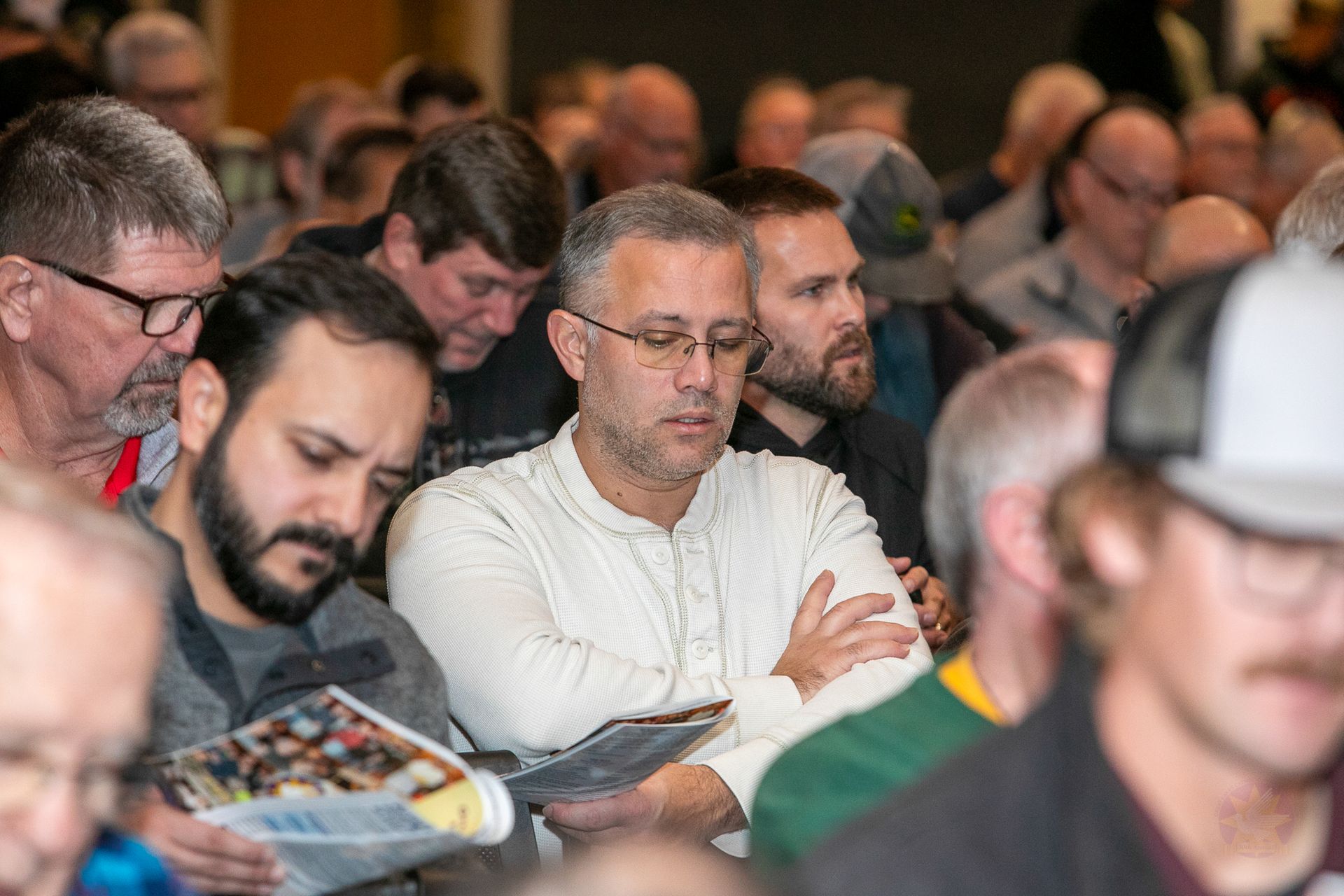 Audience seated in a conference, listening attentively with notebooks and phones in hand.