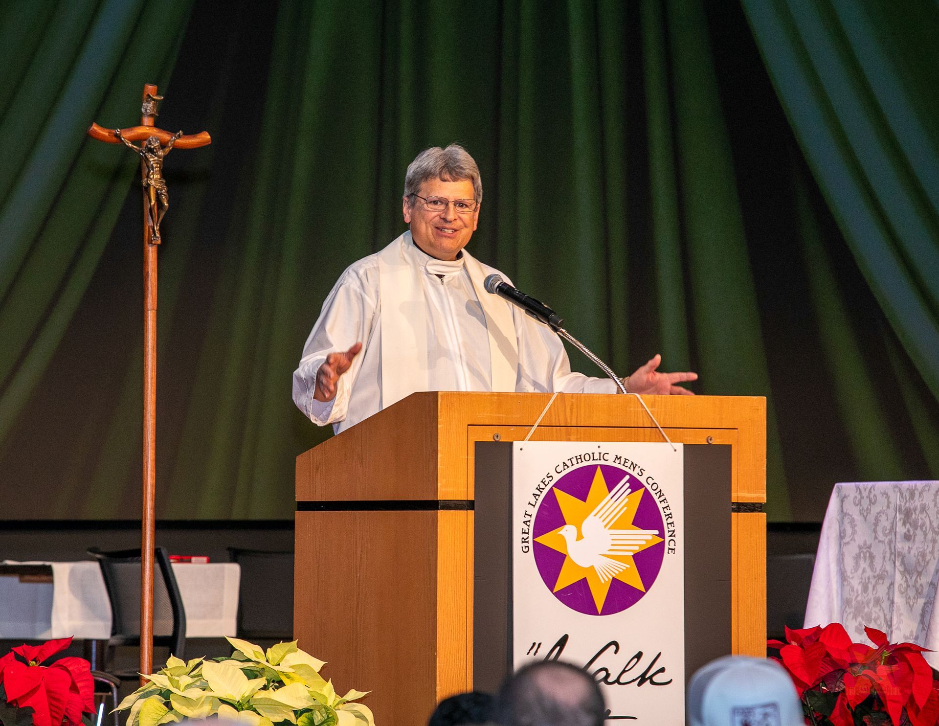 Priest at podium on stage with crucifix, dark green curtains, and flowers in front