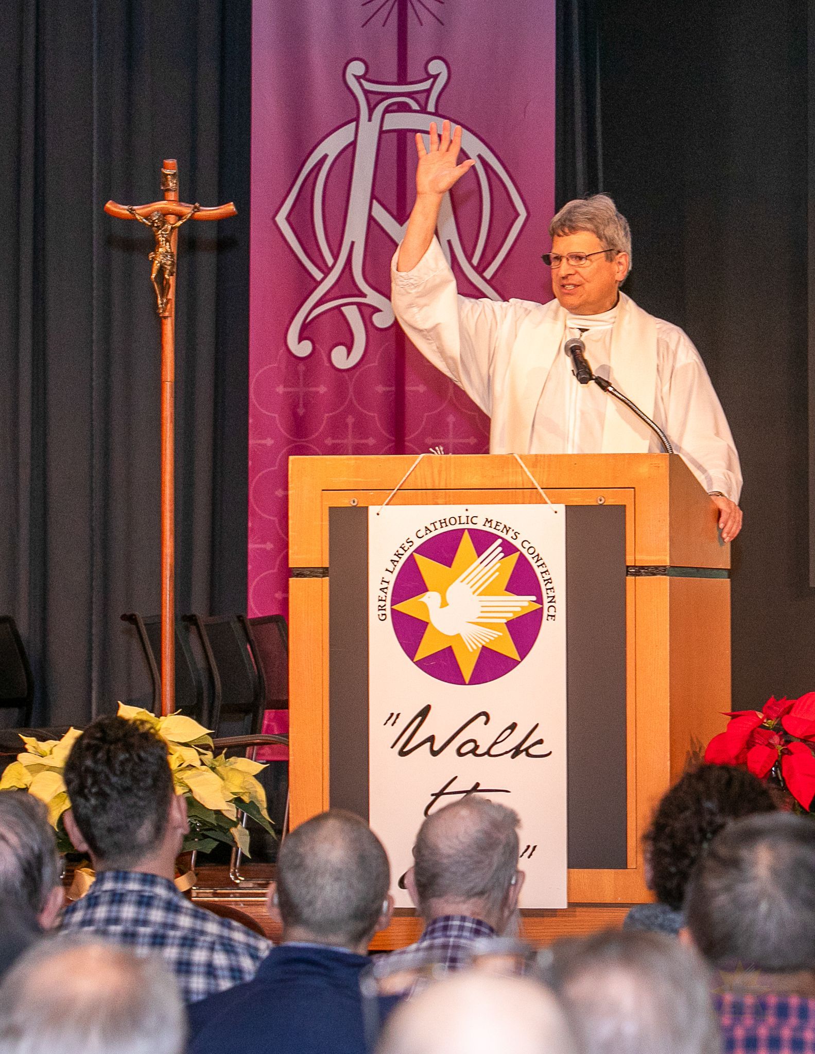 Speaker at podium raising hand during a church service, with audience seated and a cross in the background.
