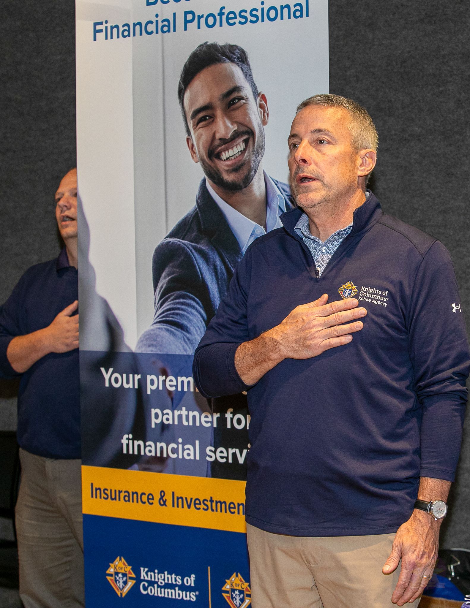 Man in navy sweater speaking beside a financial services banner at an event