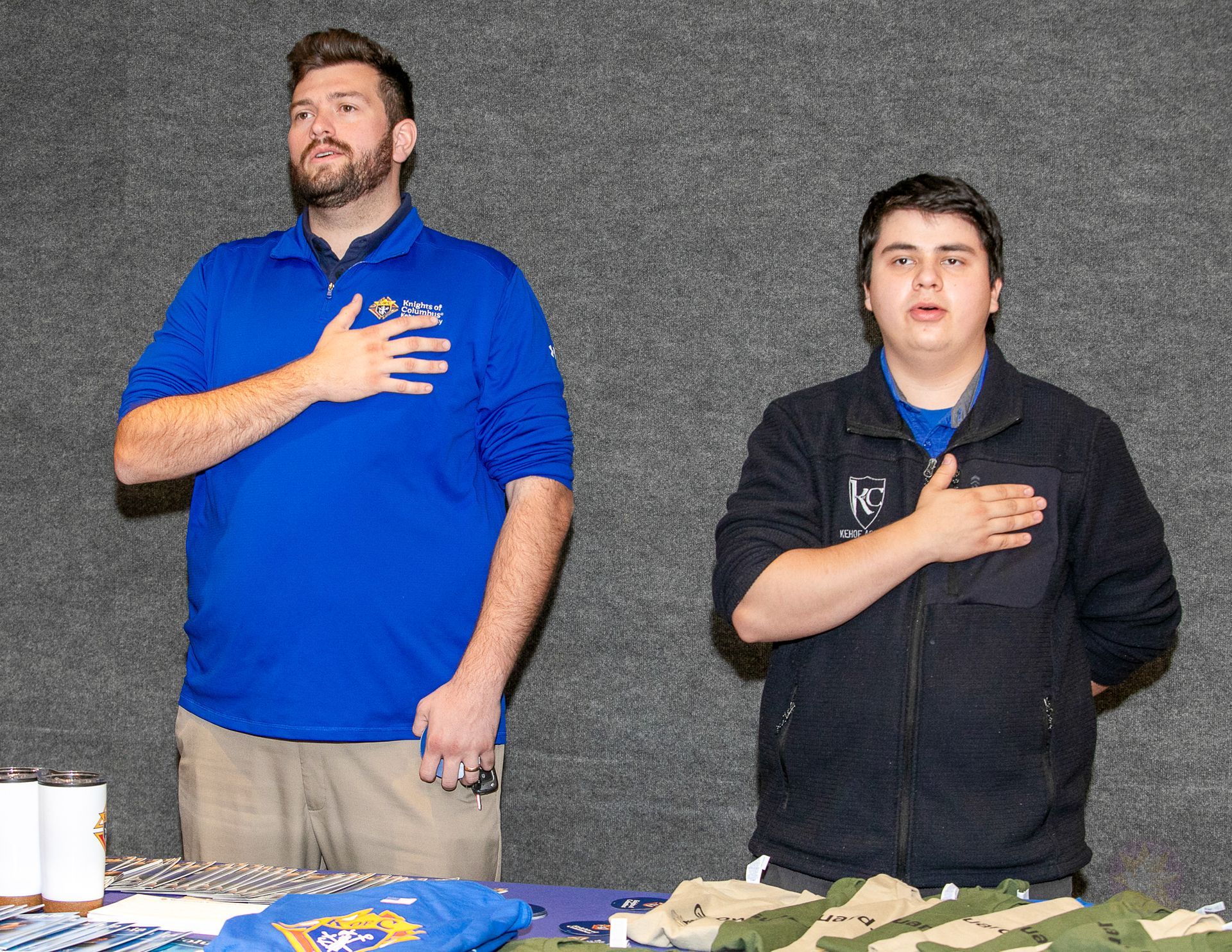 Two men standing behind a table with hands over their hearts, one in a blue shirt and one in a black jacket.