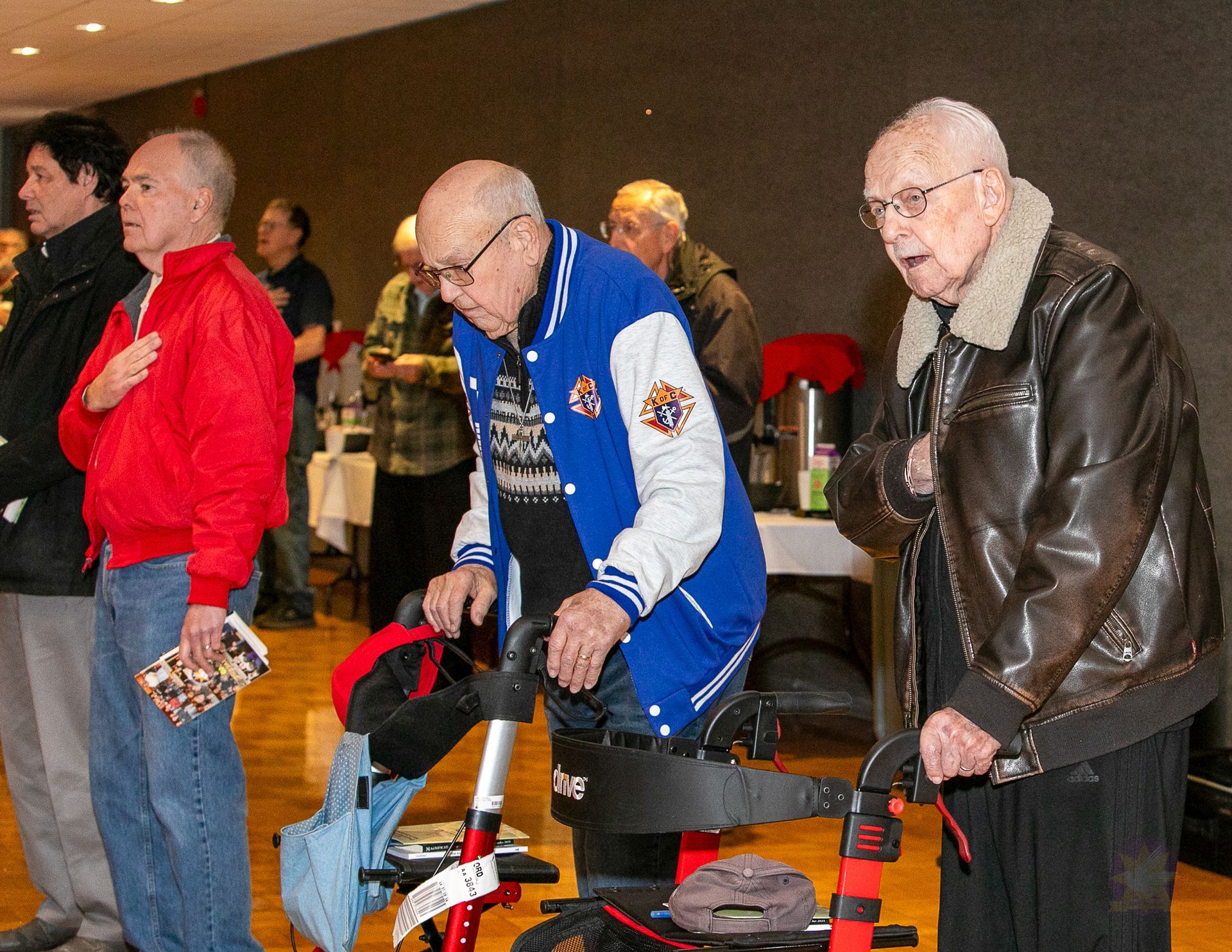 Three men standing indoors, one using a walker, wearing red, blue, and brown jackets.