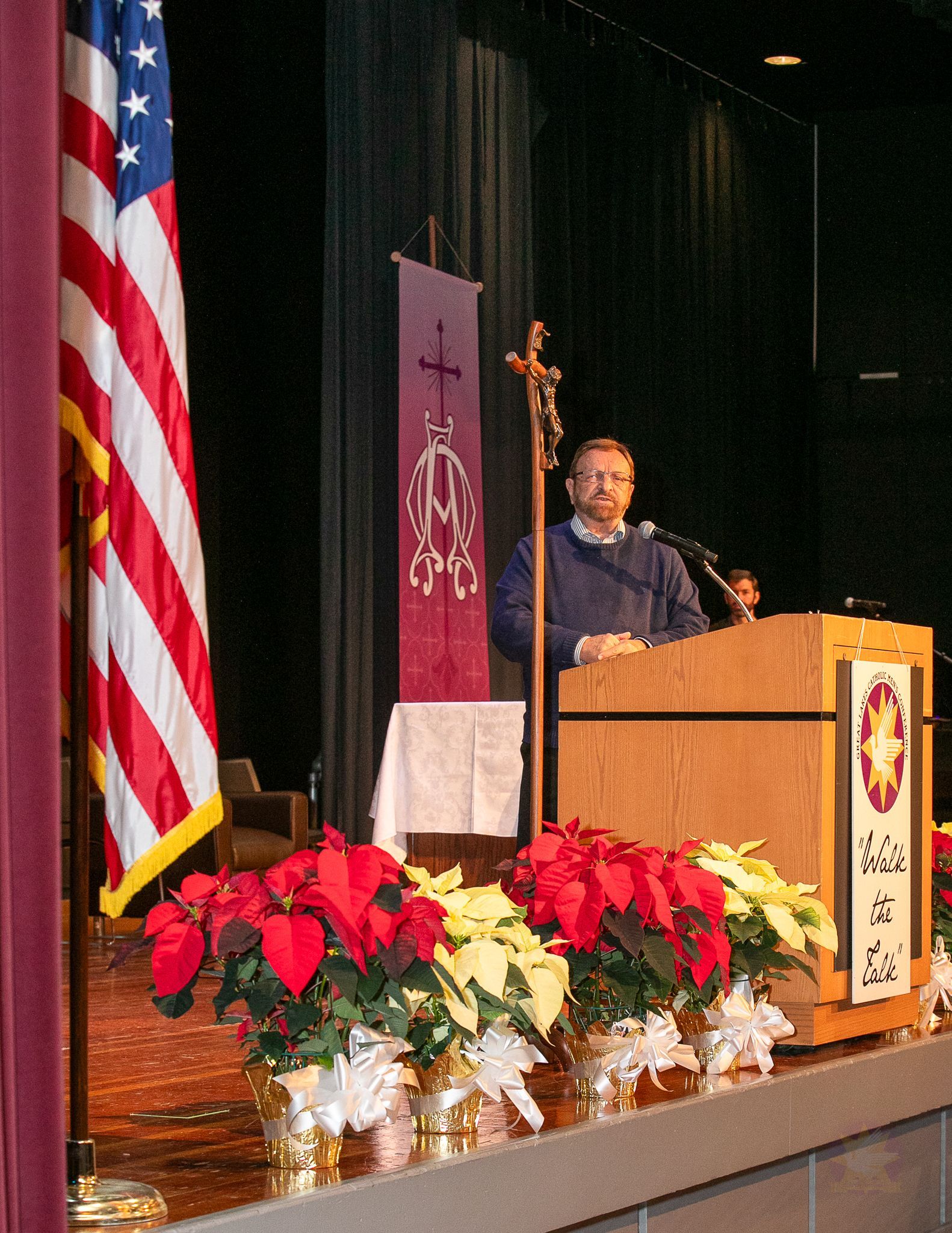 Speaker at podium on stage beside U.S. flag and poinsettias, addressing an audience.