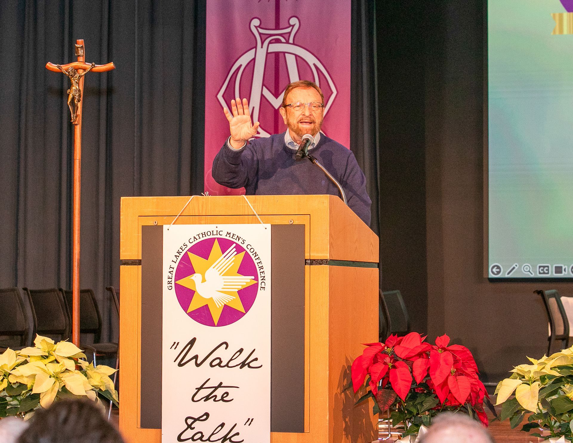 Man speaking at a podium, waving on a church stage with a cross and flowers