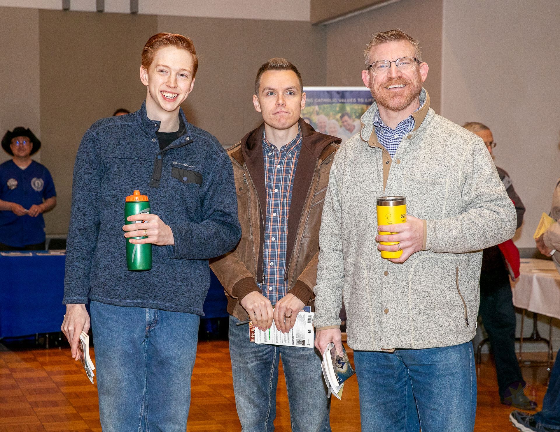 Three people standing indoors, two holding water bottles, smiling at the camera.