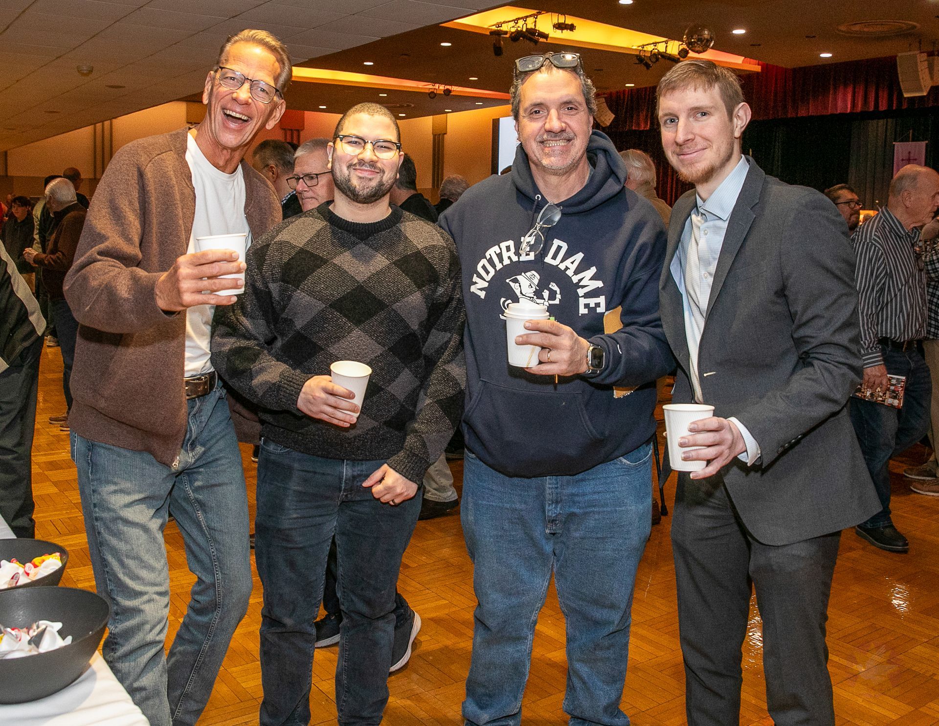 VFour people standing indoors at a Catholic men's conference, smiling and holding drinks.