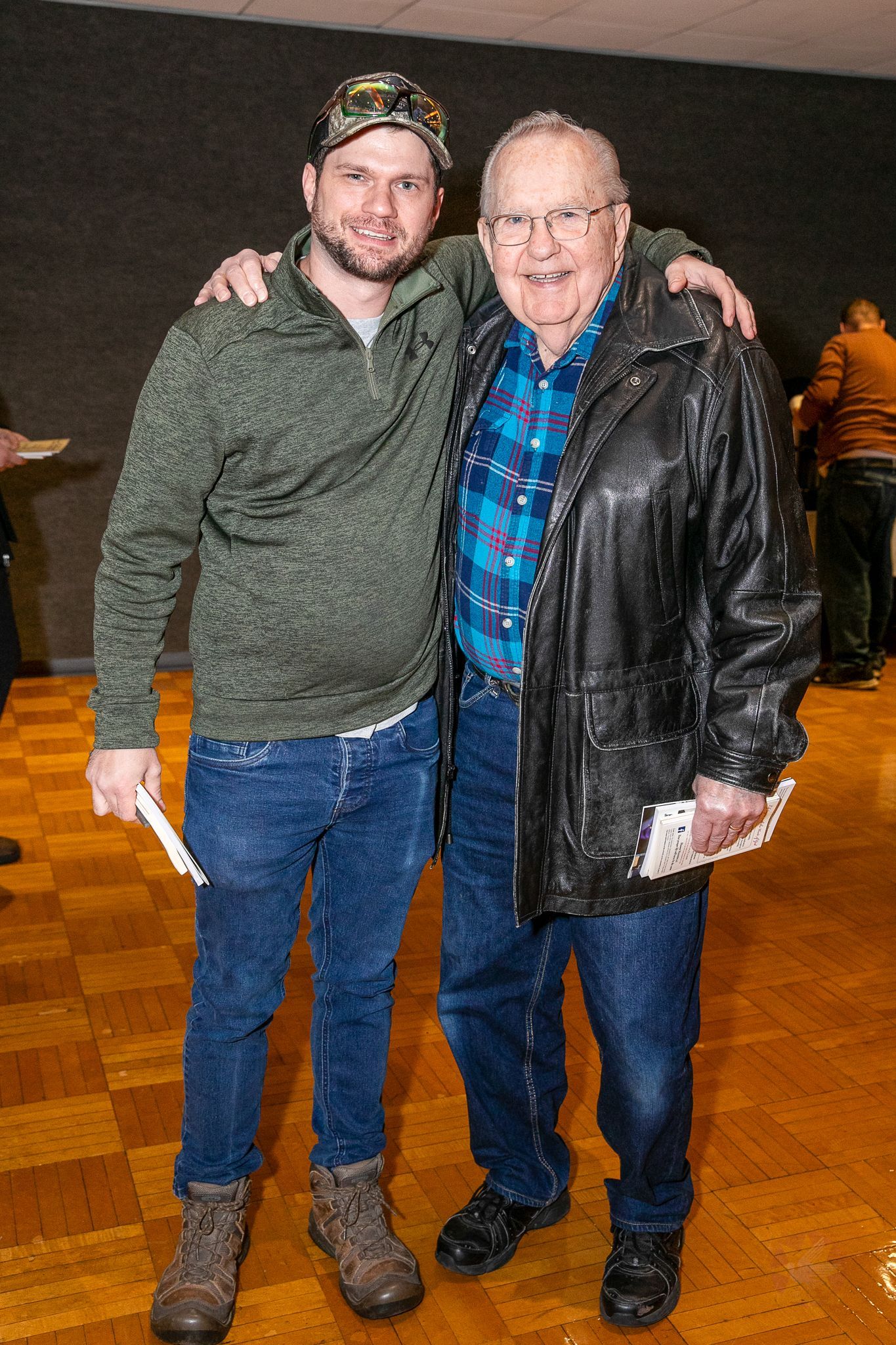 Two men smiling and posing together indoors on a wooden floor, one in a green sweater and the other in a black jacket.