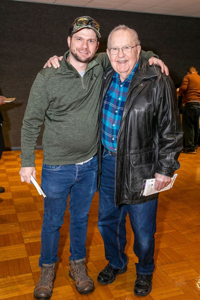 Two men smiling and posing together indoors on a wooden floor, one in a green sweater and the other in a black jacket.