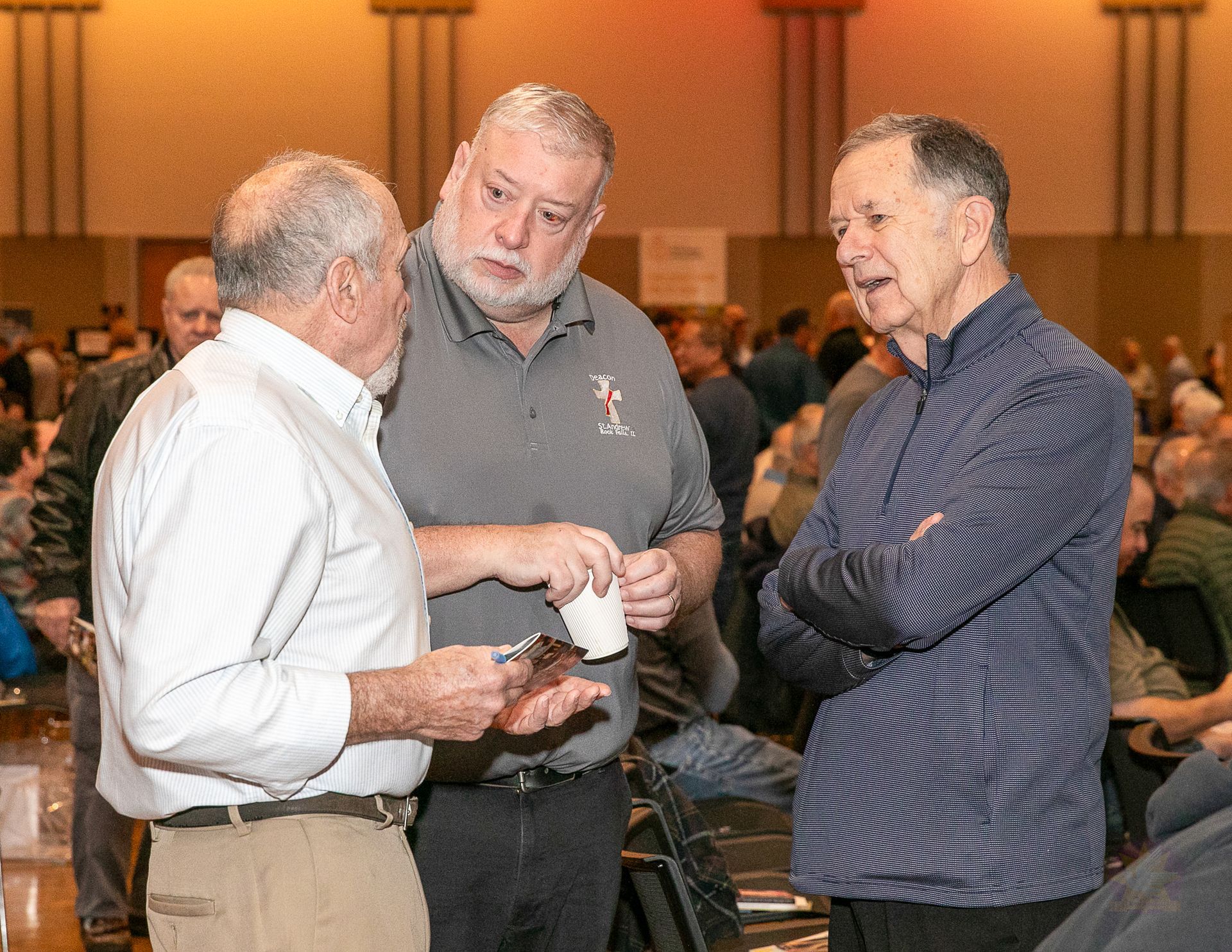Three men talking at a conference, one holding a phone, in a crowded meeting room.