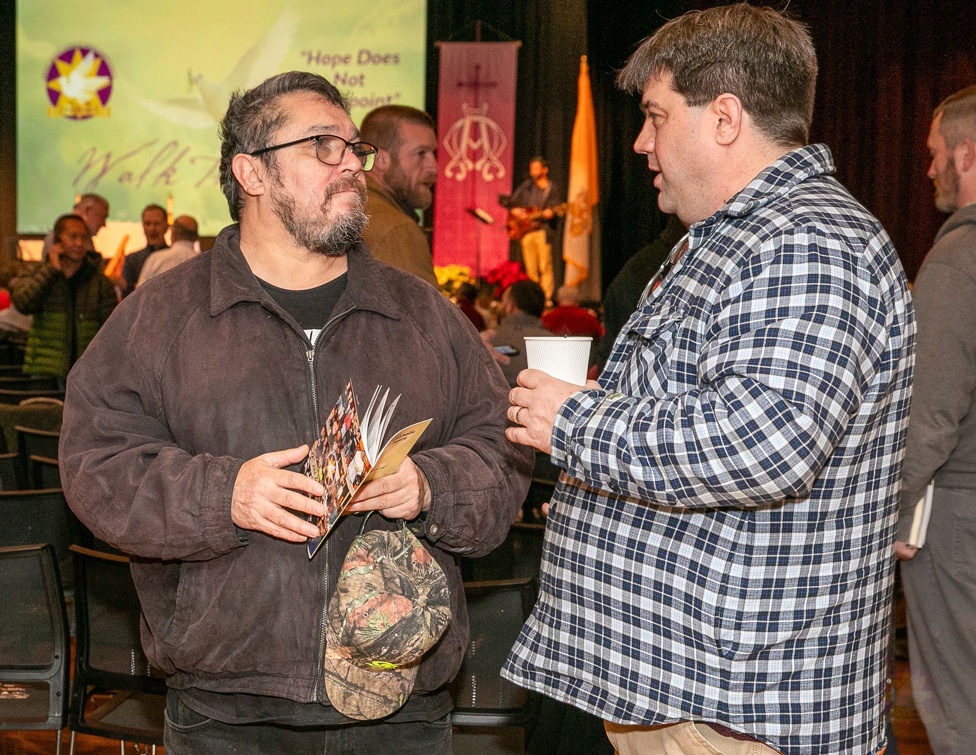 Two men chatting at an indoor event, one holding a cup and the other a clear bag with snacks.
