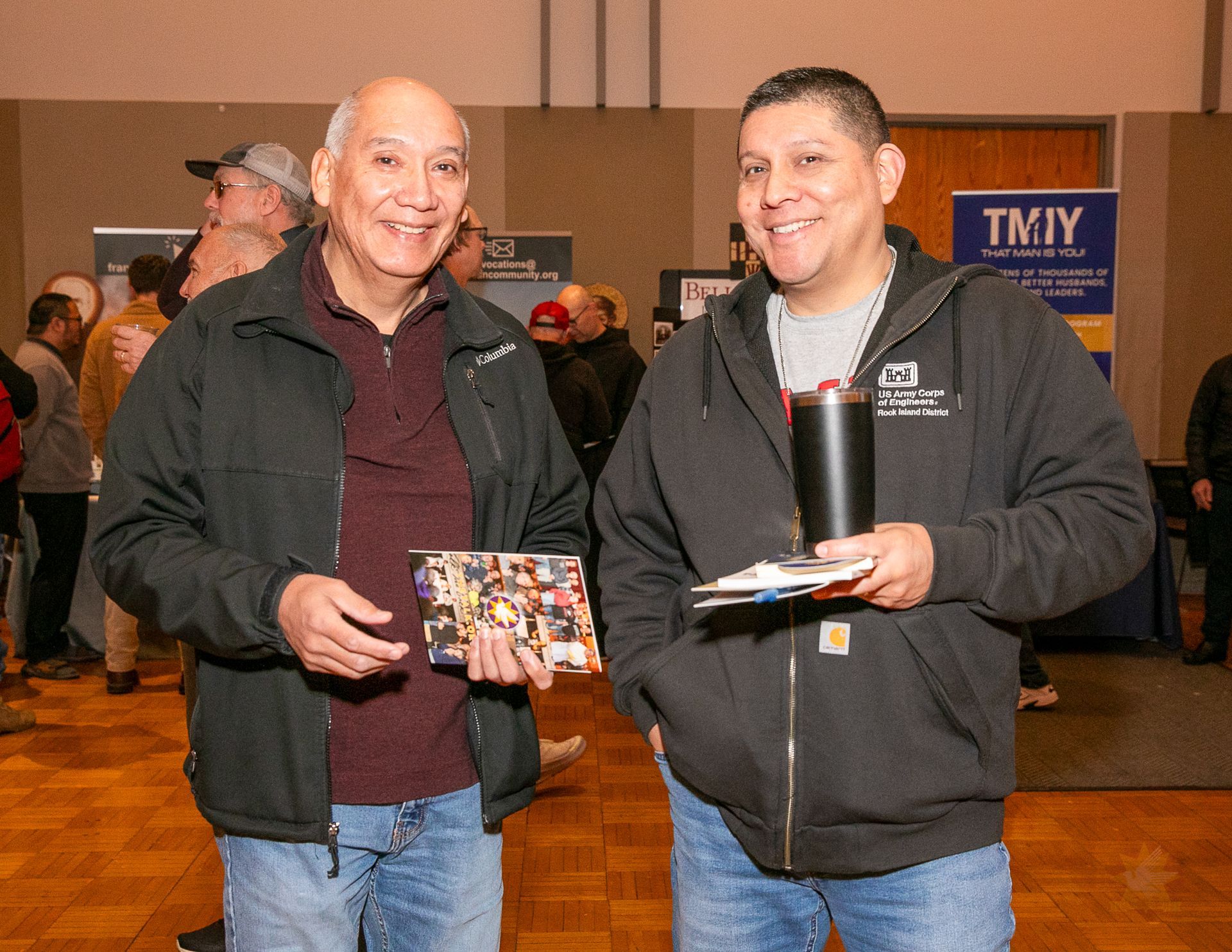 Two men smiling at an indoor event, one holding a photo and the other a black tumbler and plate.