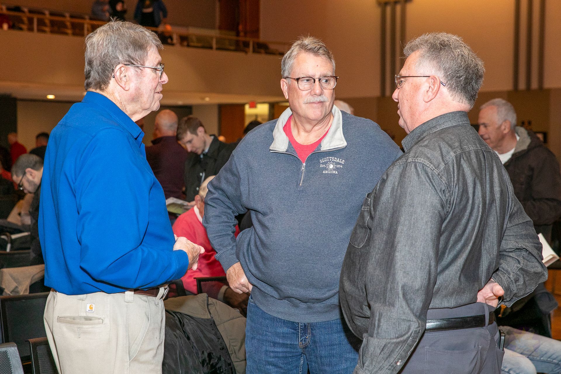 Three men chatting at an indoor event, one in a blue shirt, one in a gray sweater, one in a black jacket.