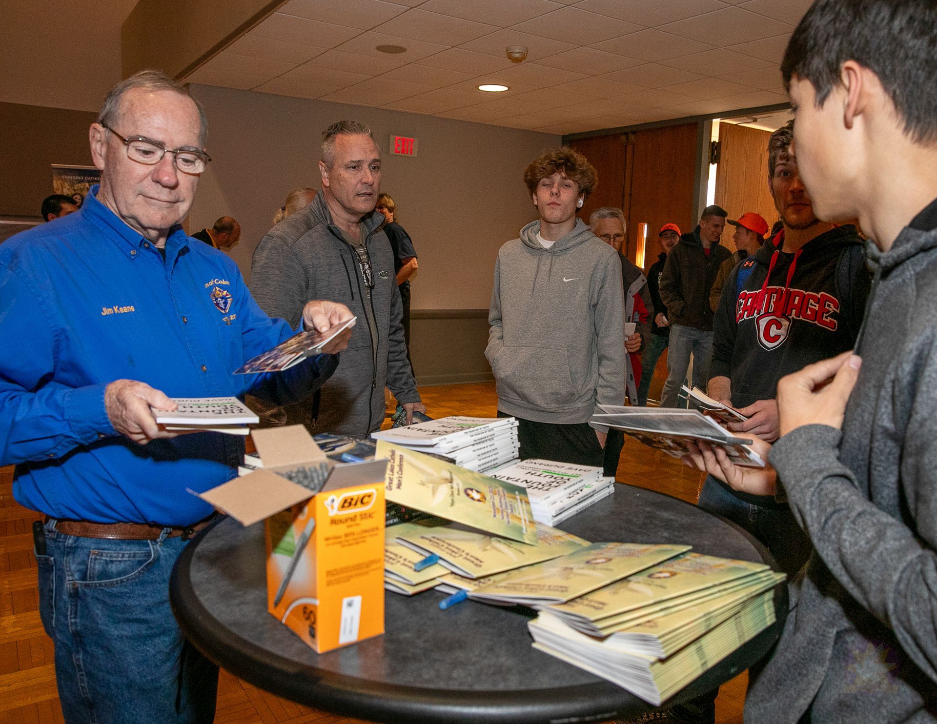 People exchanging flyers at a table at a Catholic men's conference, with stacks of brochures and a box of snacks.
