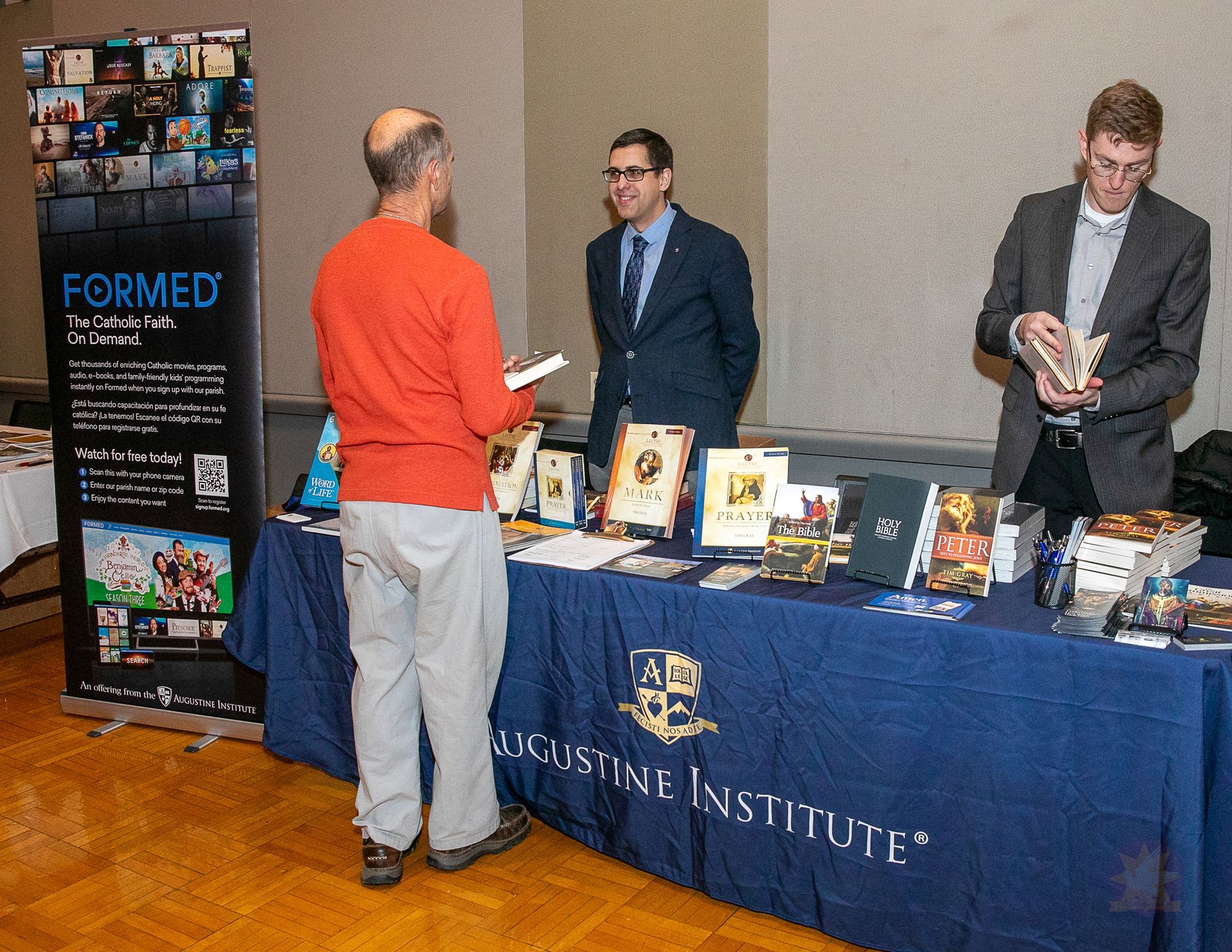 Book fair booth with table displays and three people talking in a conference hall.