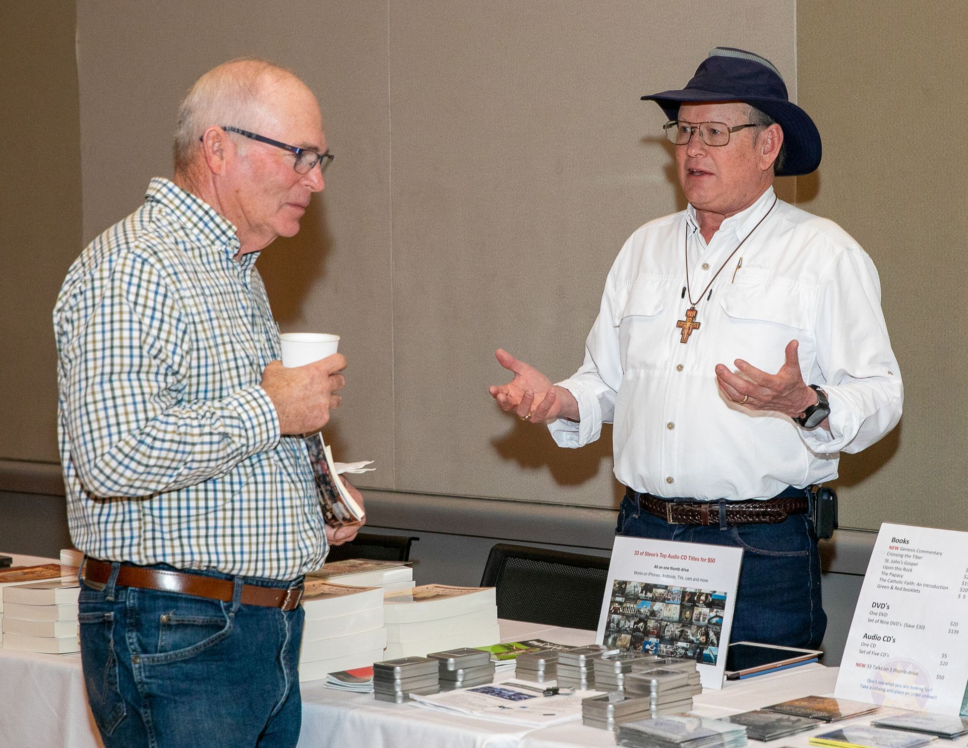 Two men conversing at a display table with brochures, one holding a cup and the other gesturing while wearing a hat.