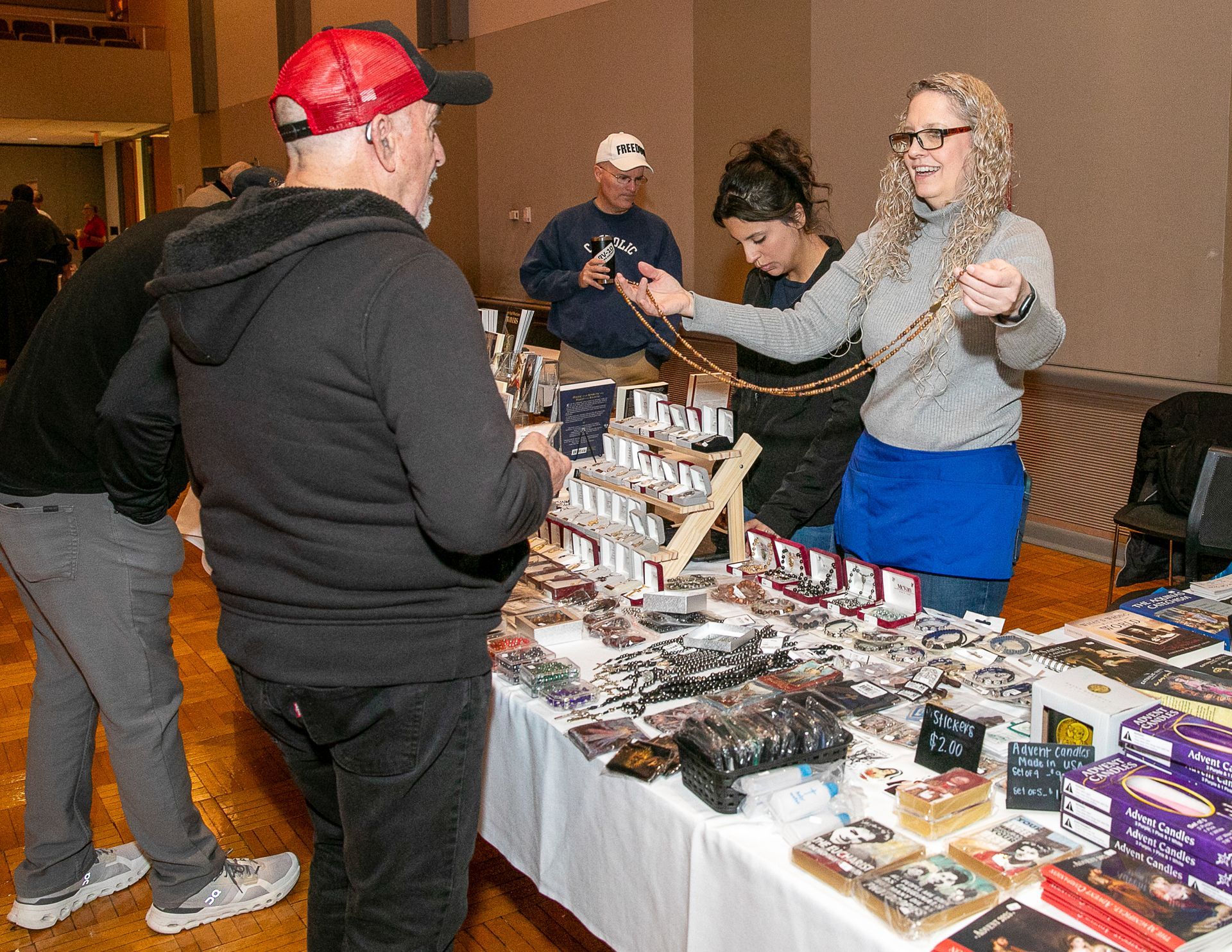 Vendor booth with people browsing a table of books, crafts, and merchandise at an indoor event.
