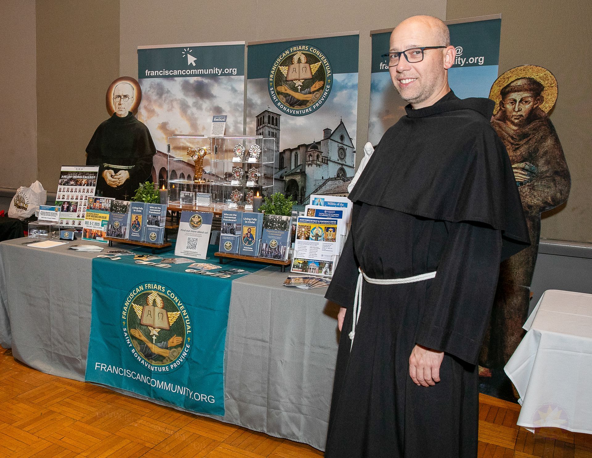 Two people in black robes standing behind a display table with religious items and banners.