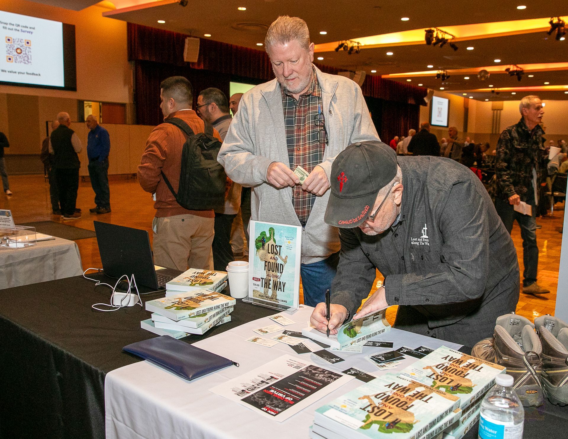 Man signing a book at a table during an indoor event, with another person standing nearby.