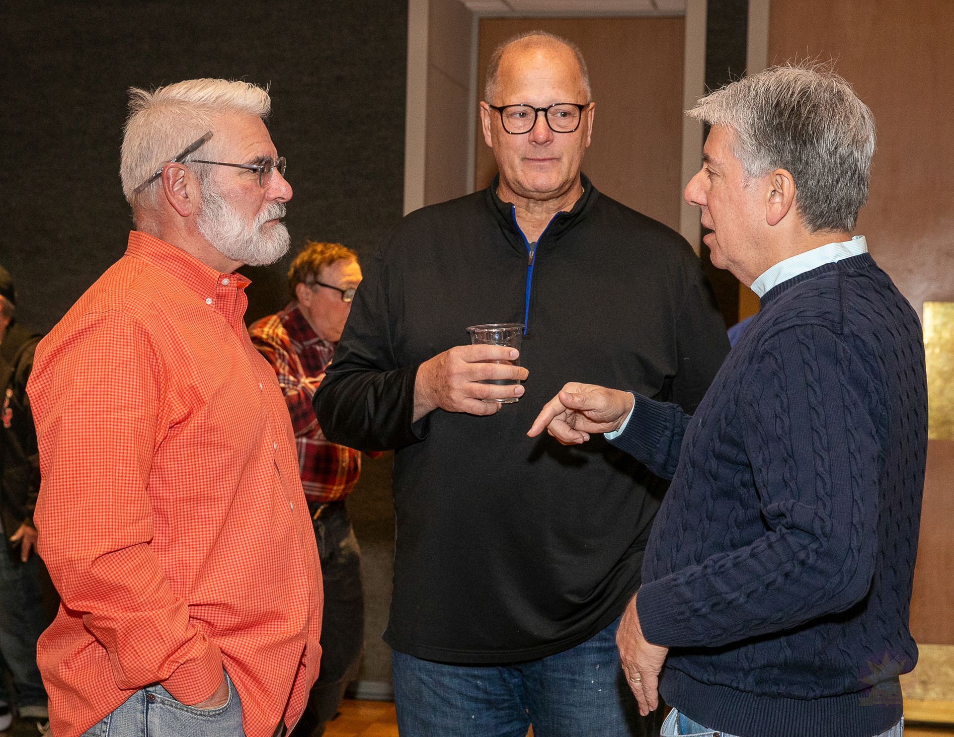 Three men in conversation indoors, one holding a drink and another wearing a bright orange shirt.