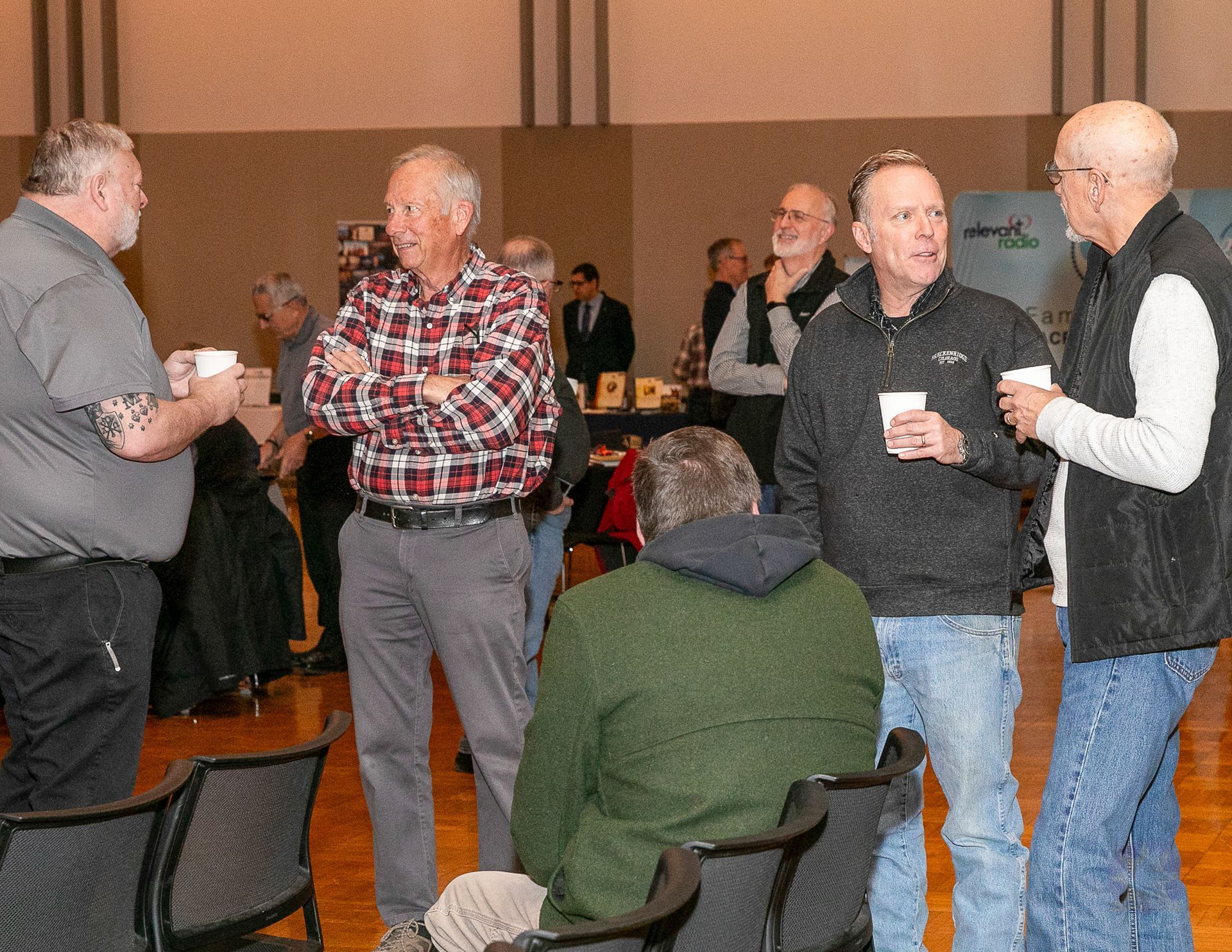Several people chat at a casual indoor gathering, some holding drinks and wearing jackets and plaid shirts.