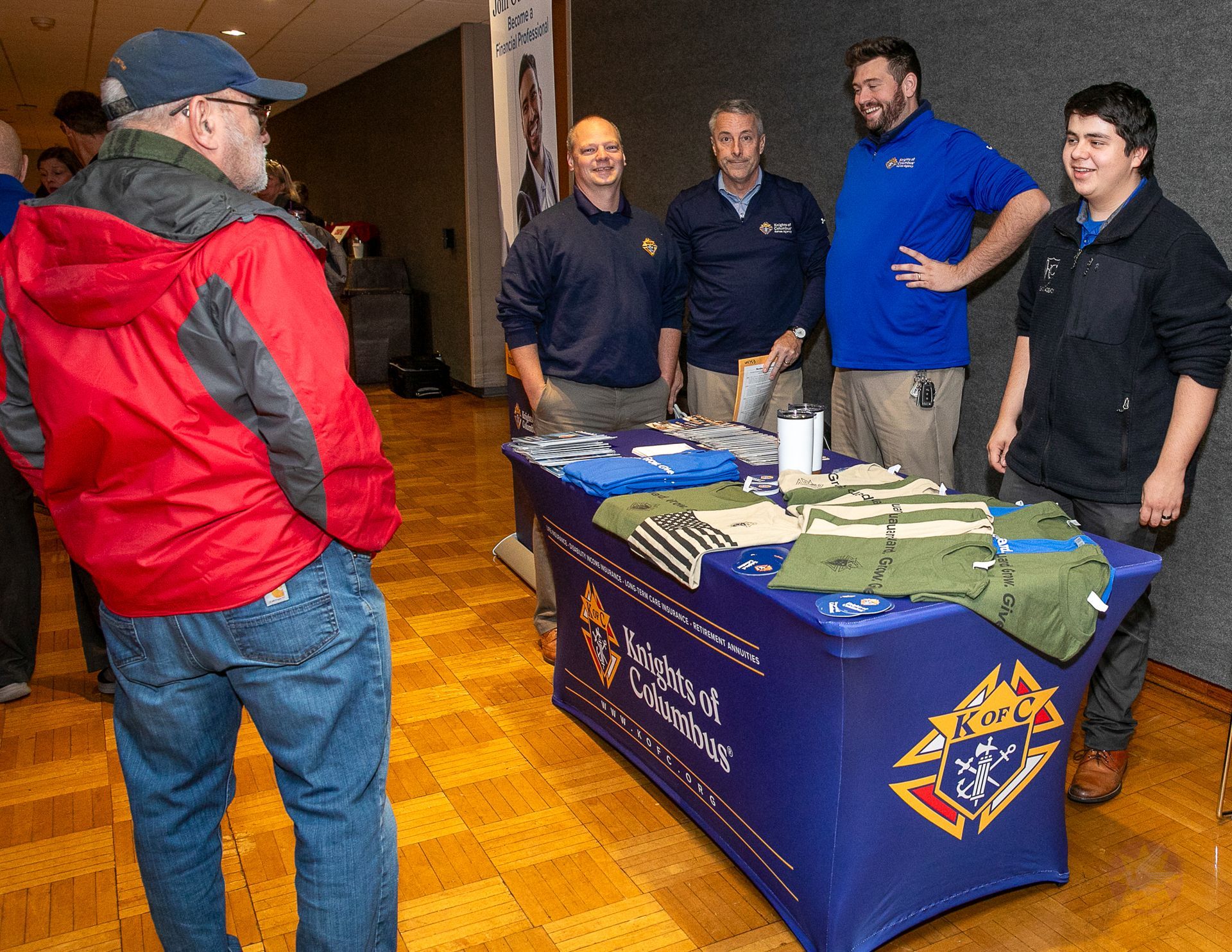 Three men behind a blue table at an indoor event, greeting a man in a red jacket.