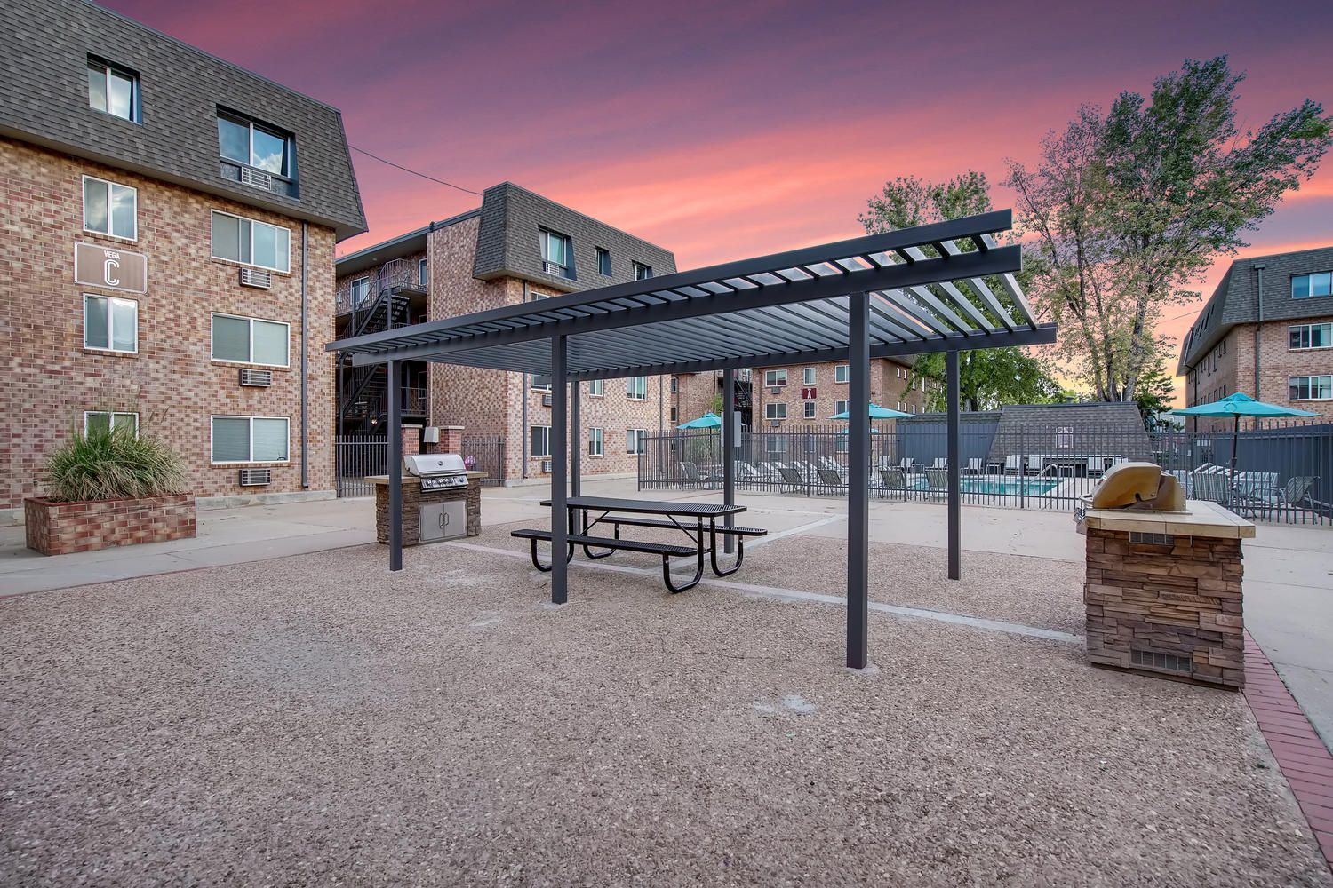 Outdoor seating area with picnic table under a pergola, grilling station, and apartment buildings at dusk.