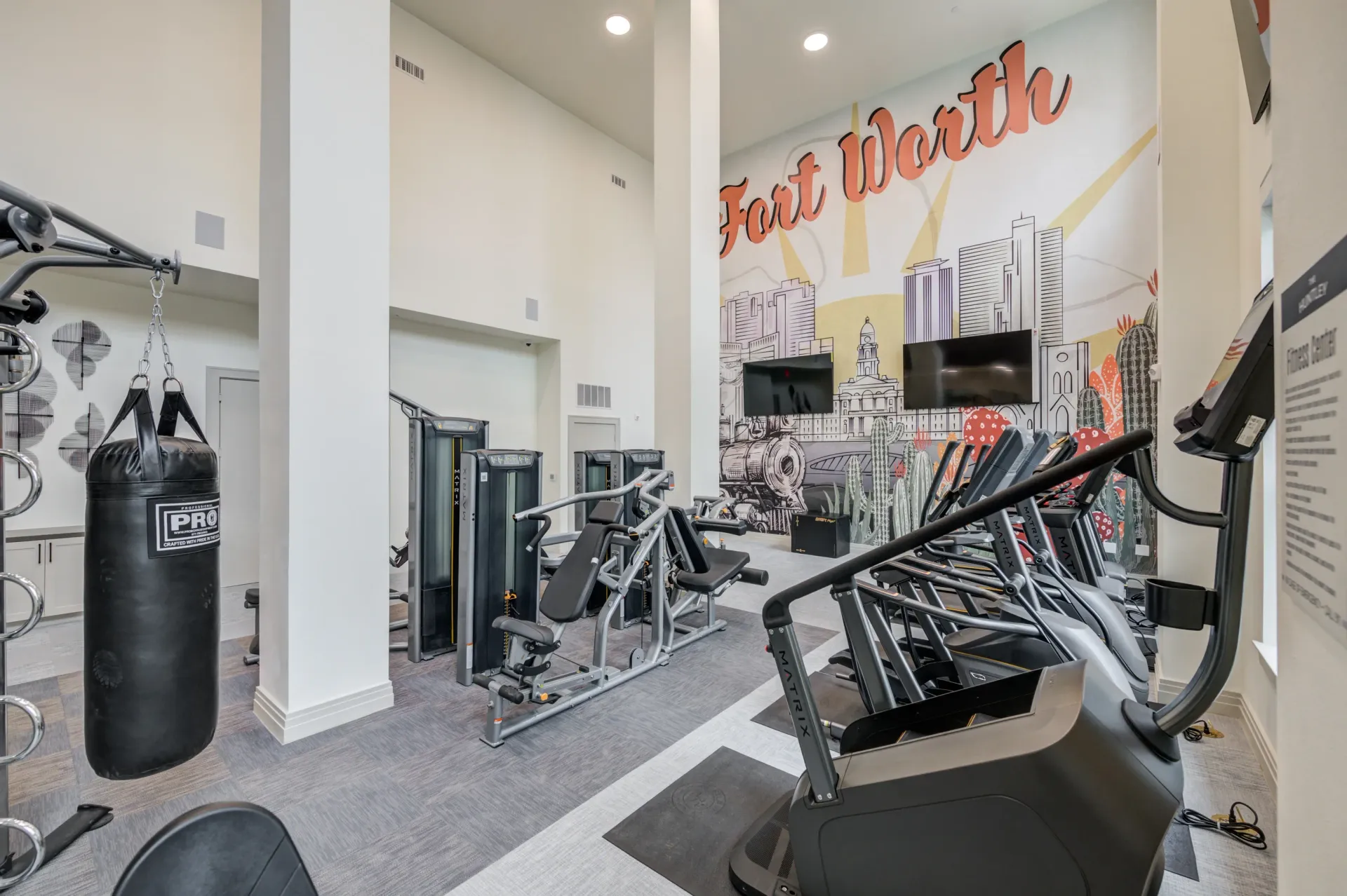 Gym with cardio equipment, weight machines, and a punching bag, featuring a Fort Worth mural.
