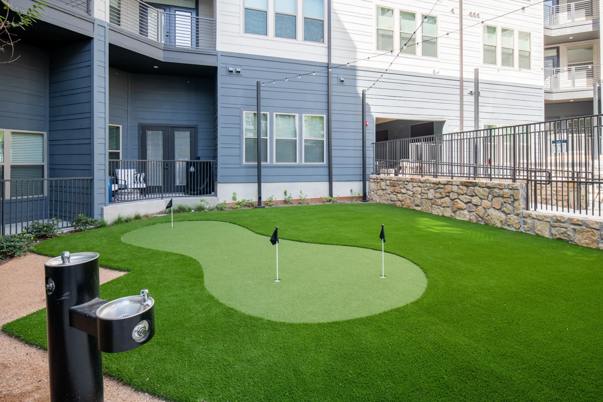 Putting green with flags and a drinking fountain outside apartment buildings.