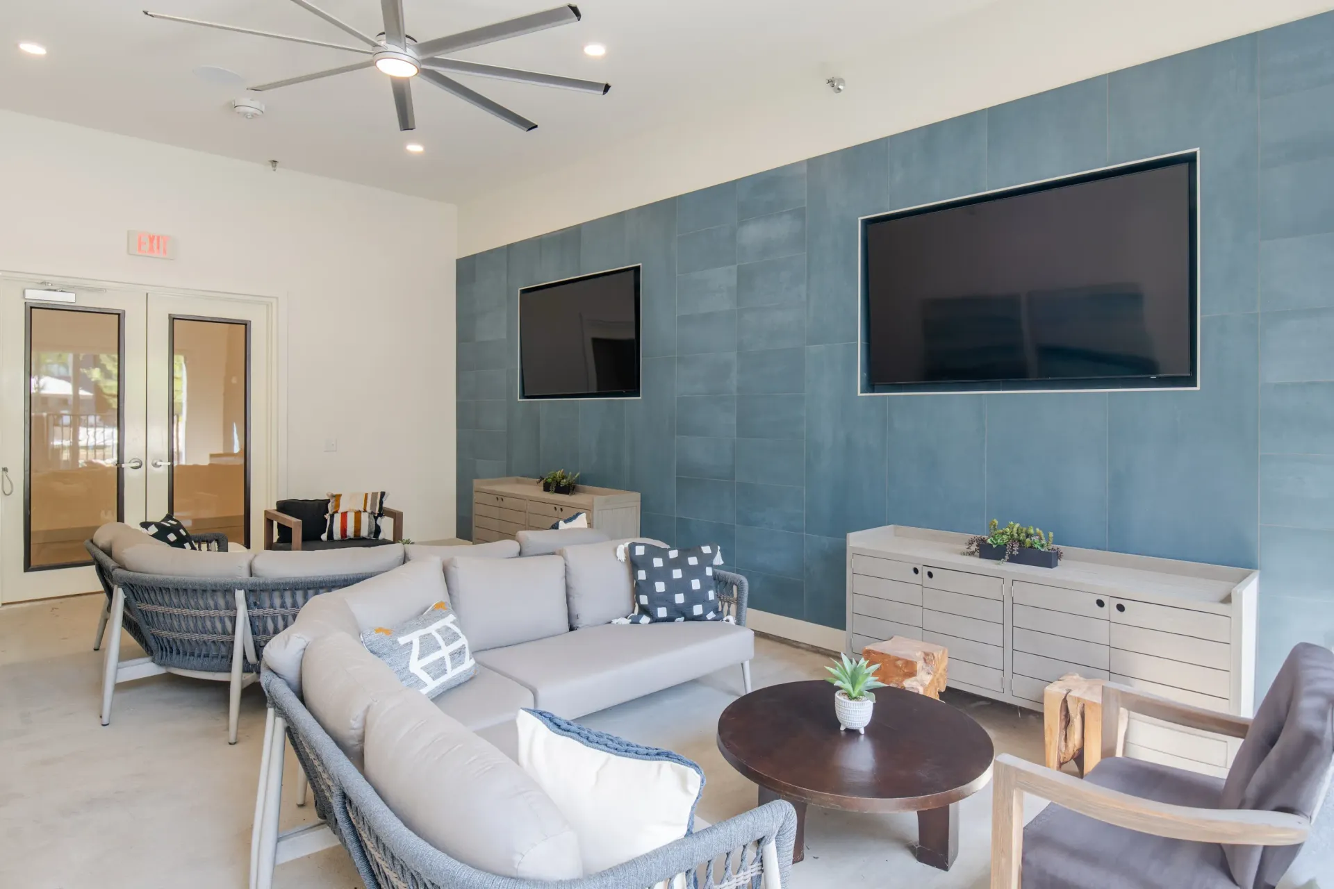 Communal lounge area with sectional sofa, chairs, and two televisions mounted on a blue tiled wall.