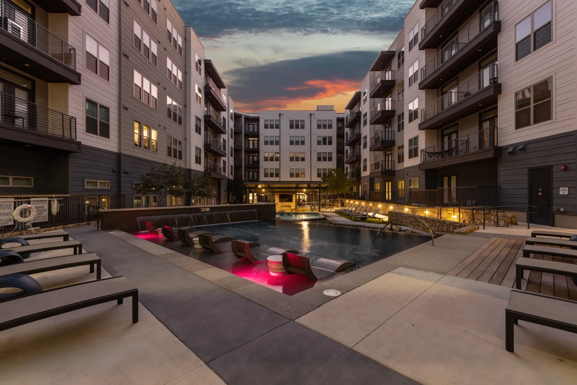 Modern apartment complex courtyard with a swimming pool and lounge chairs at twilight.