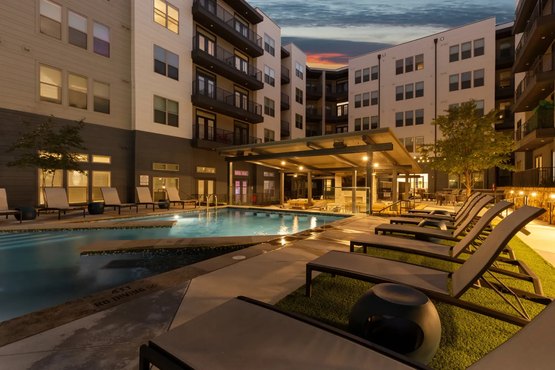 Modern apartment building exterior with a swimming pool and lounge chairs at dusk.