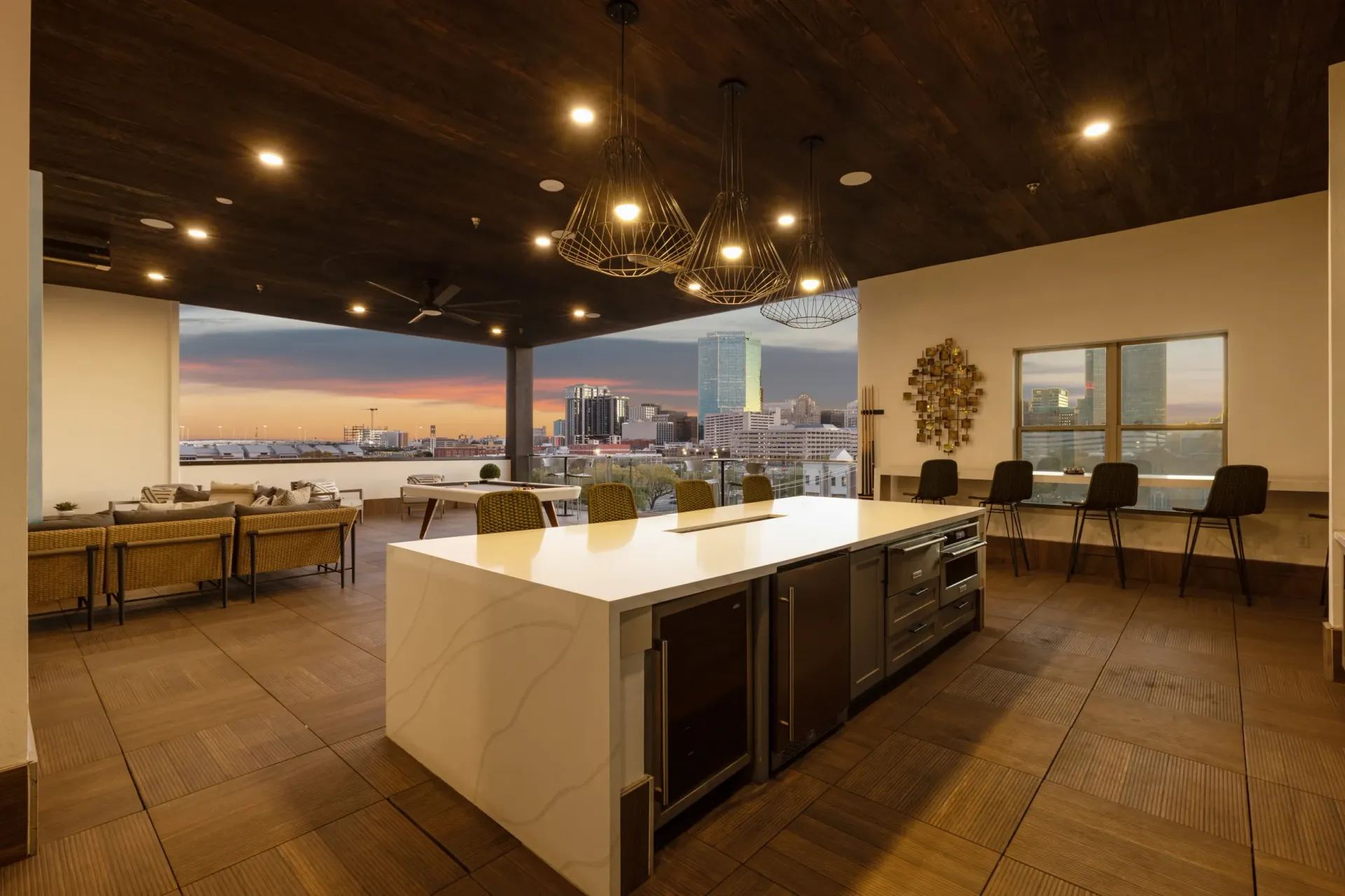 Modern rooftop lounge with seating, a pool table, and a large island kitchen overlooking the city skyline at sunset.
