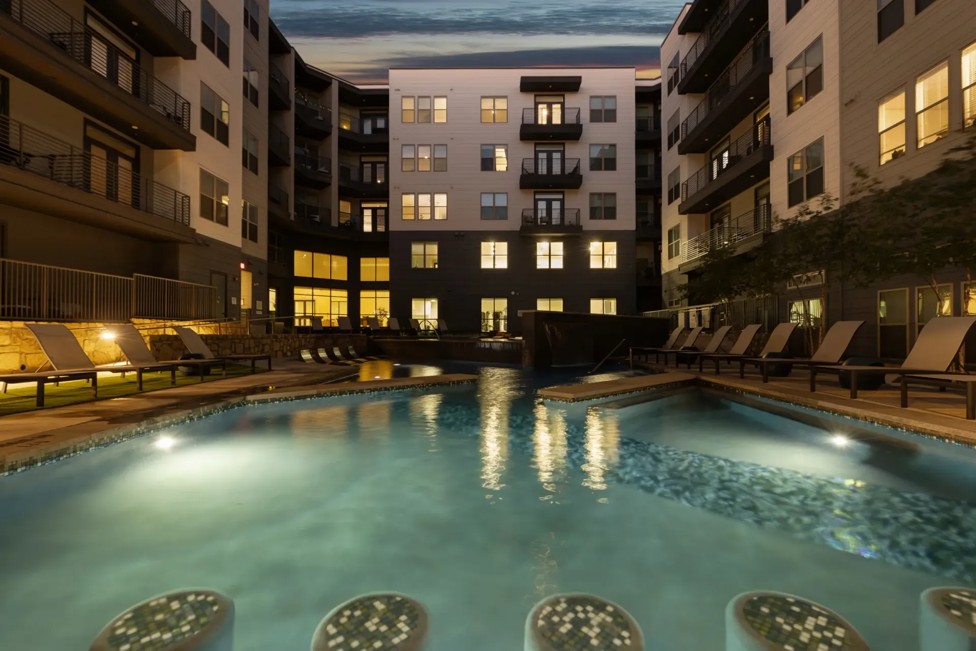 Modern apartment complex courtyard with a swimming pool and lounge chairs at dusk.