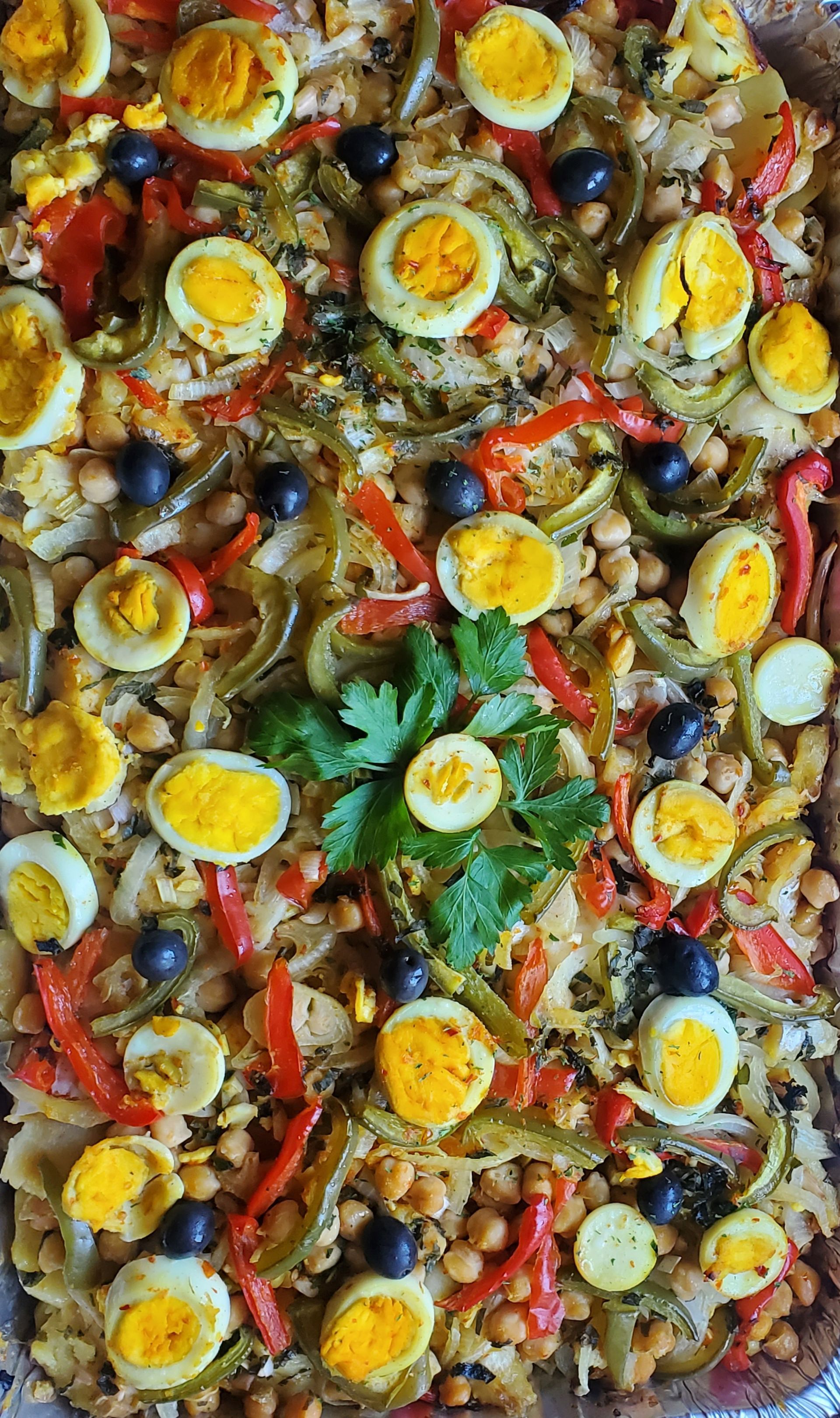 Overhead shot of a vibrant, cooked dish with halved eggs, olives, peppers, and chickpeas.