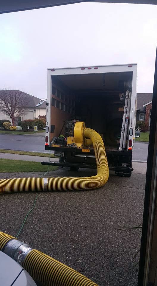 Yellow blower with hose filling a truck's cargo area, parked in a driveway.