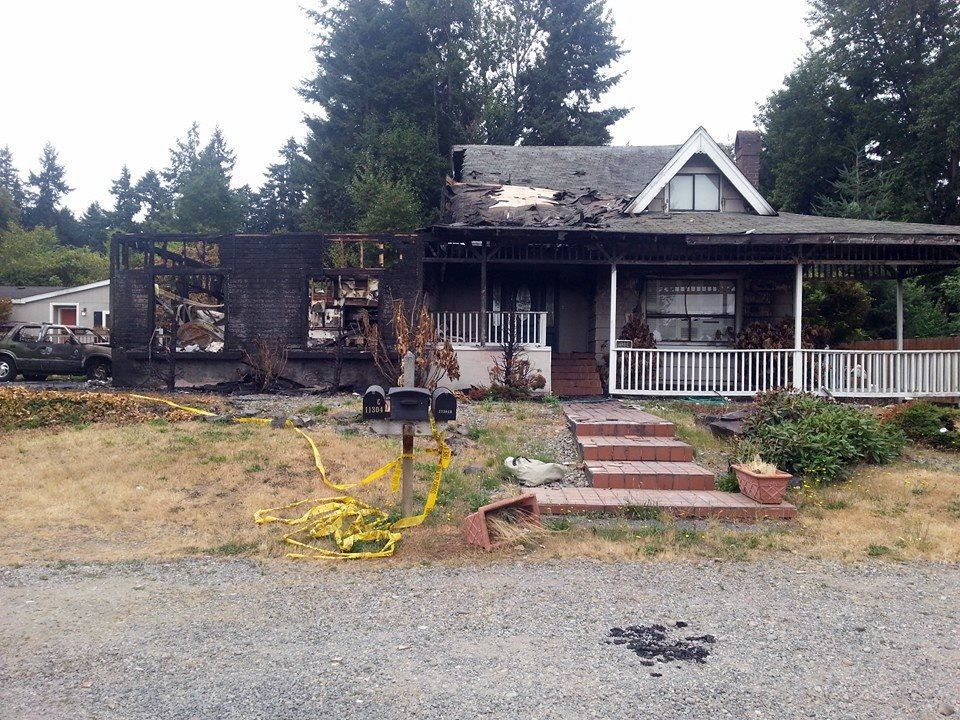 House with charred remains from a fire; yellow caution tape in yard; overcast sky.